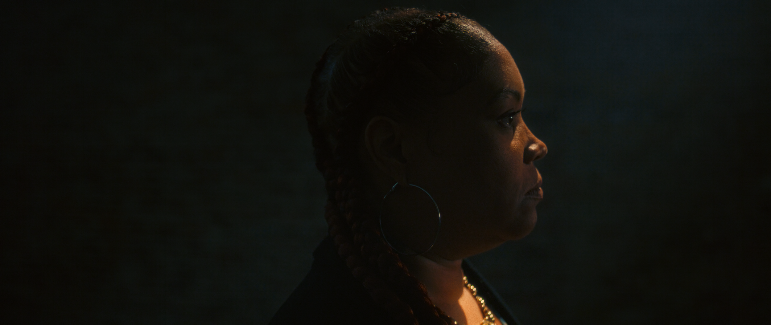 Side profile of a woman with braided hair and large hoop earrings, illuminated softly from the front, against a dark background.