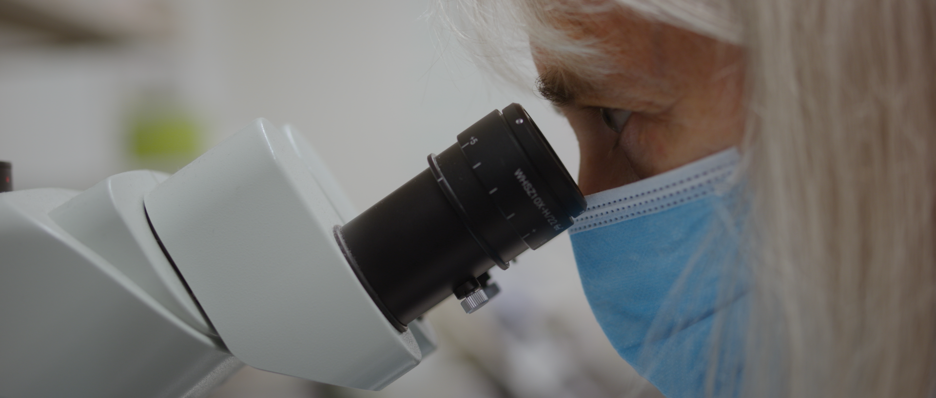 A scientist wearing a blue mask looking through a microscope in a laboratory.
