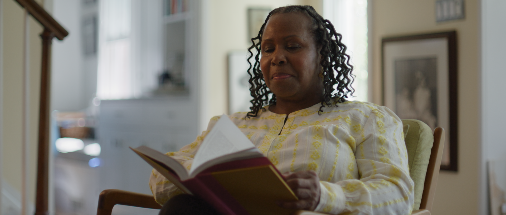 An older woman with curly hair sits in a green armchair, reading a book in a cozy, well-lit room with framed pictures on the wall and a blurred background.