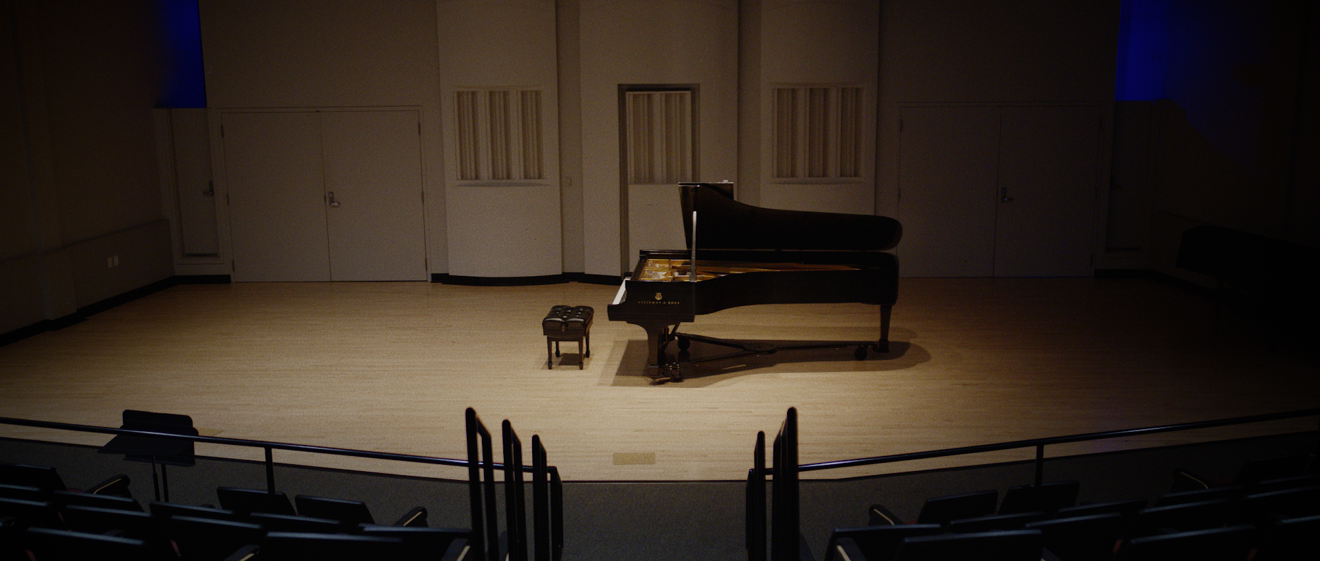 Empty concert hall stage with a grand piano and a small bench on stage, with rows of auditorium seats in the foreground.