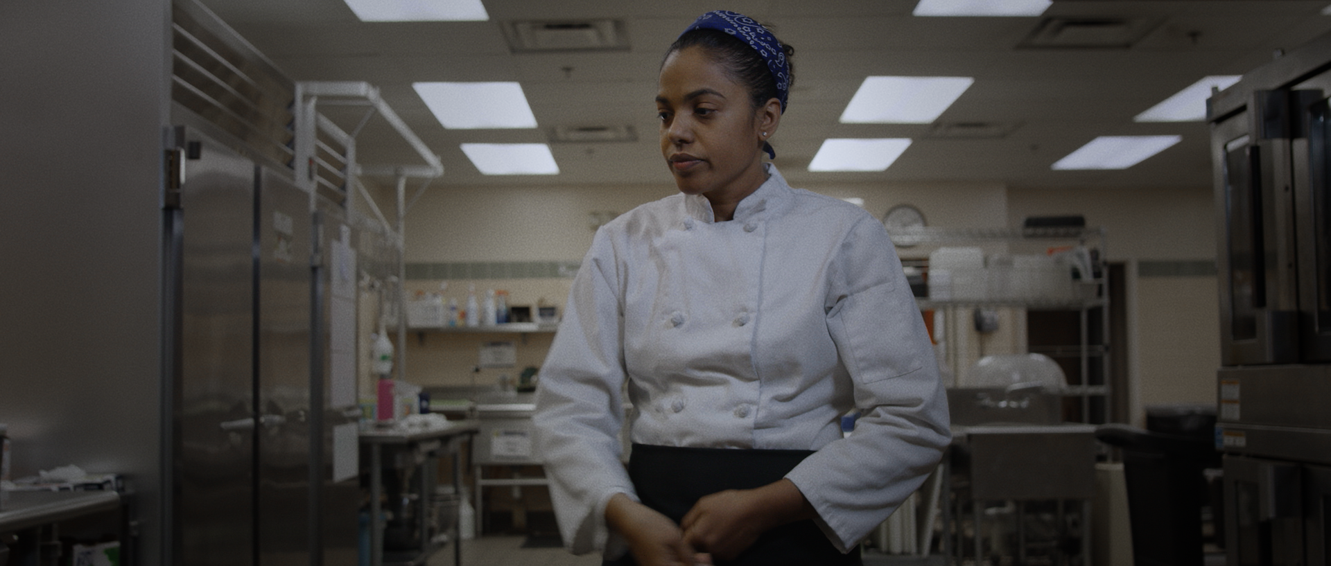 A female chef in a white chef's coat and blue bandana in a commercial kitchen.