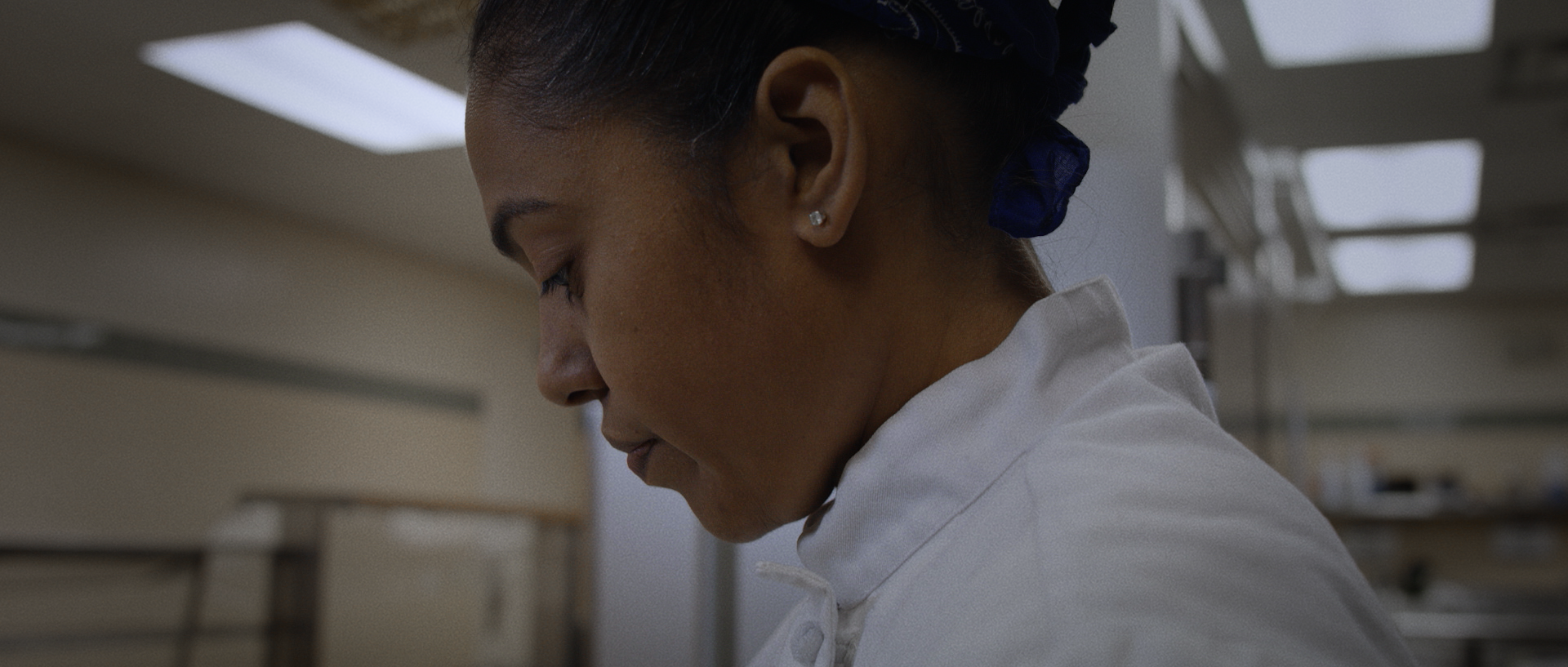 Close-up of a woman in a white coat with a blue hair bonnet in a clinical setting, with her eyes closed and head slightly bowed.