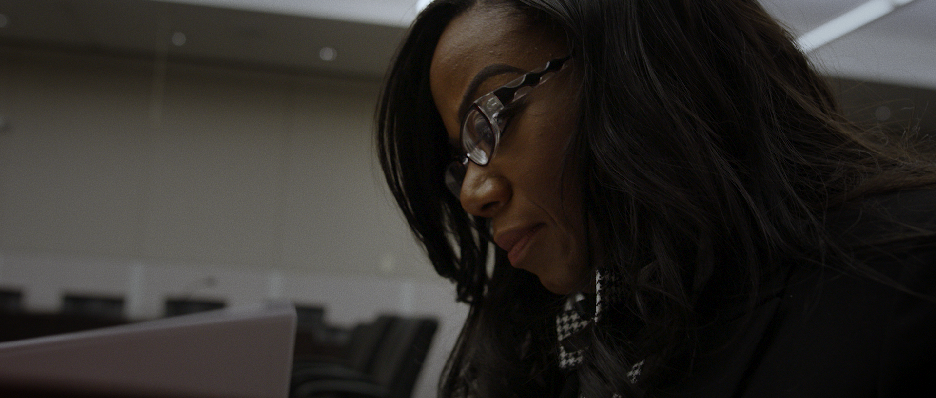 A woman with dark hair and glasses looking downward in a professional setting.