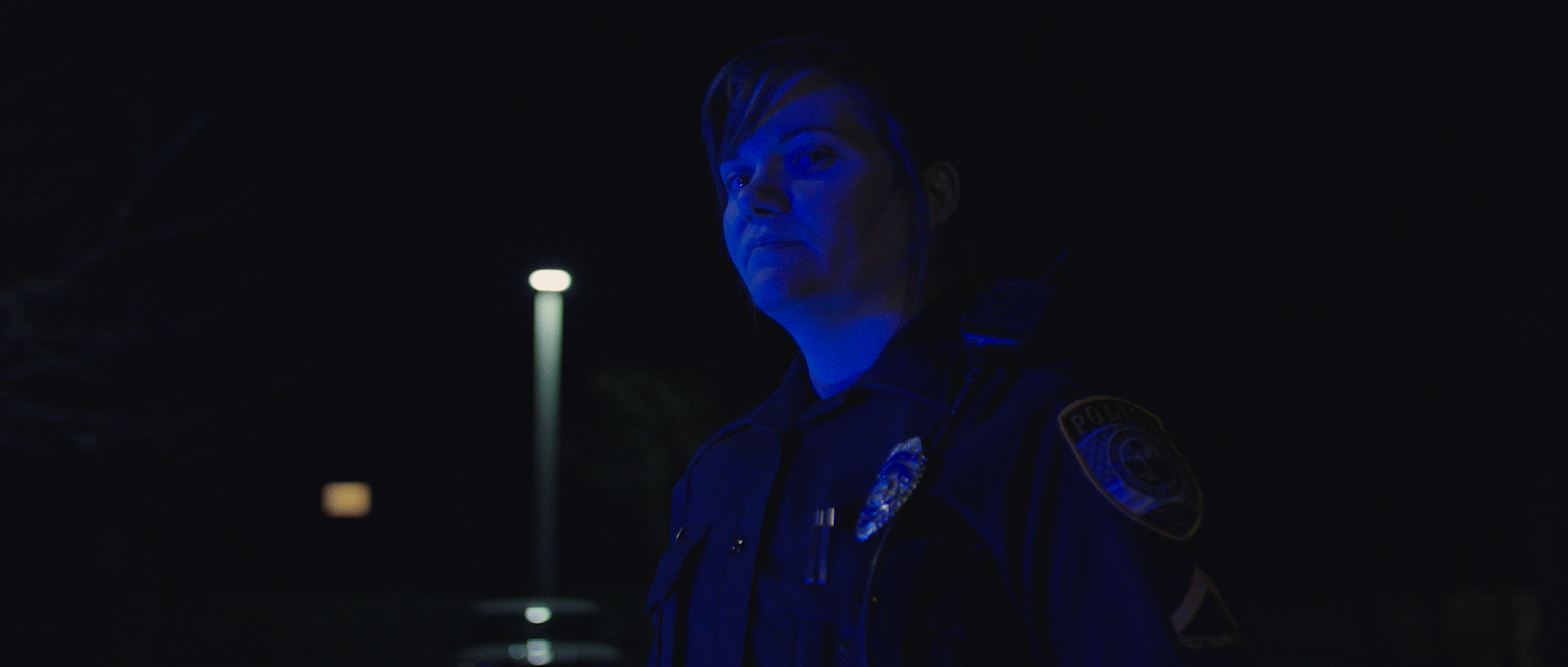 Nighttime scene featuring a police officer illuminated by blue light, standing outdoors with a streetlamp and trees in the background.