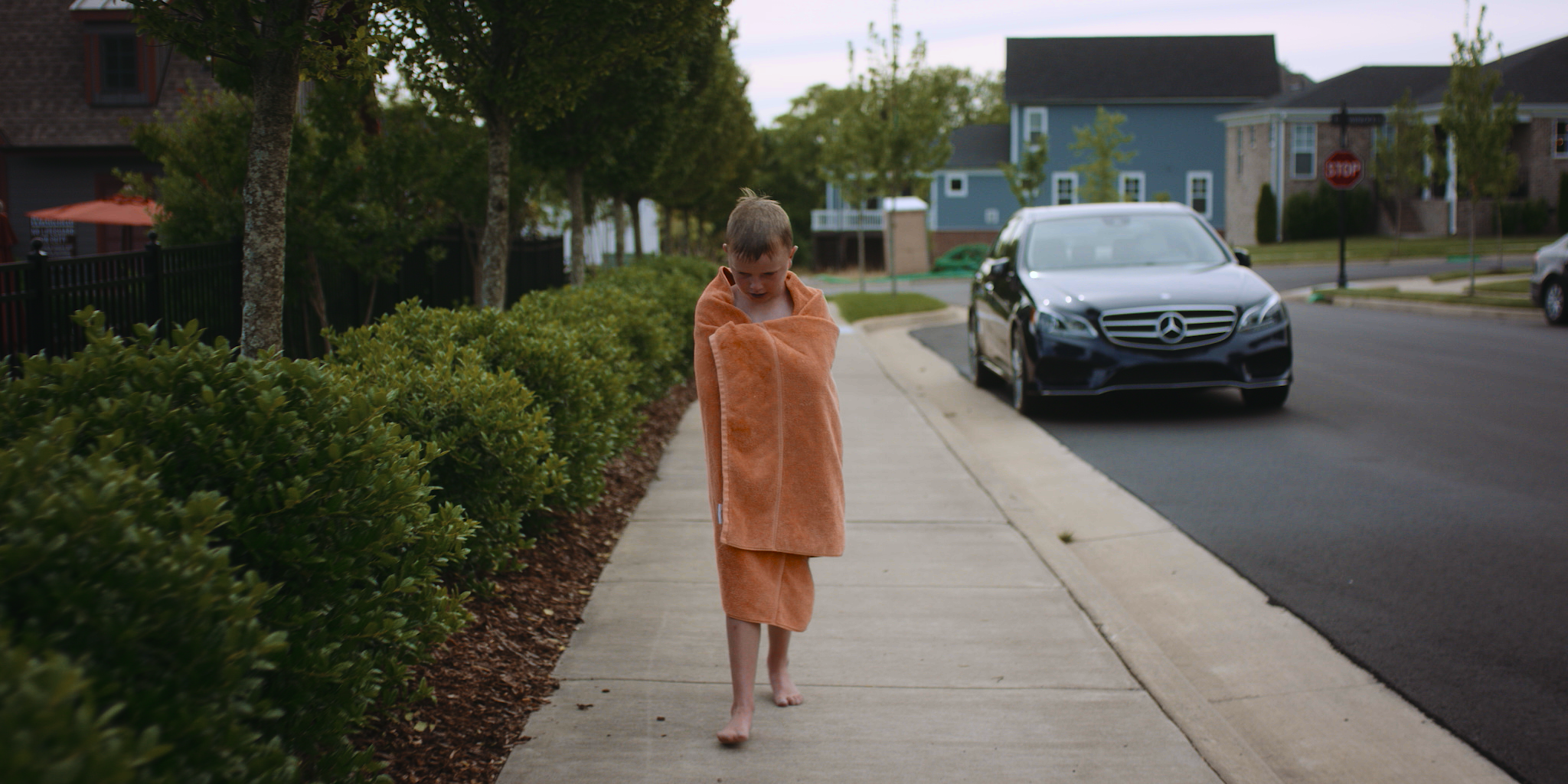 A young boy wrapped in an orange towel walking barefoot on a sidewalk in a residential neighborhood with cars parked along the street and houses in the background.