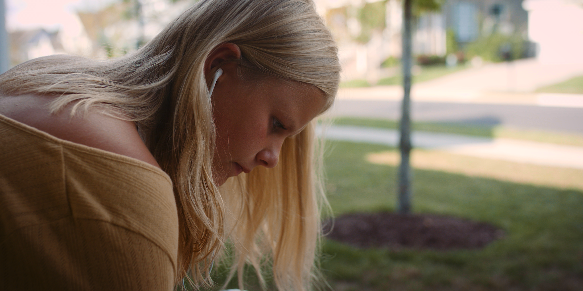 A young girl with long blonde hair and light skin, looking downwards, wearing white earbuds and a tan sweater, sitting outside near a grassy area and trees, with houses and a street in the blurred background.