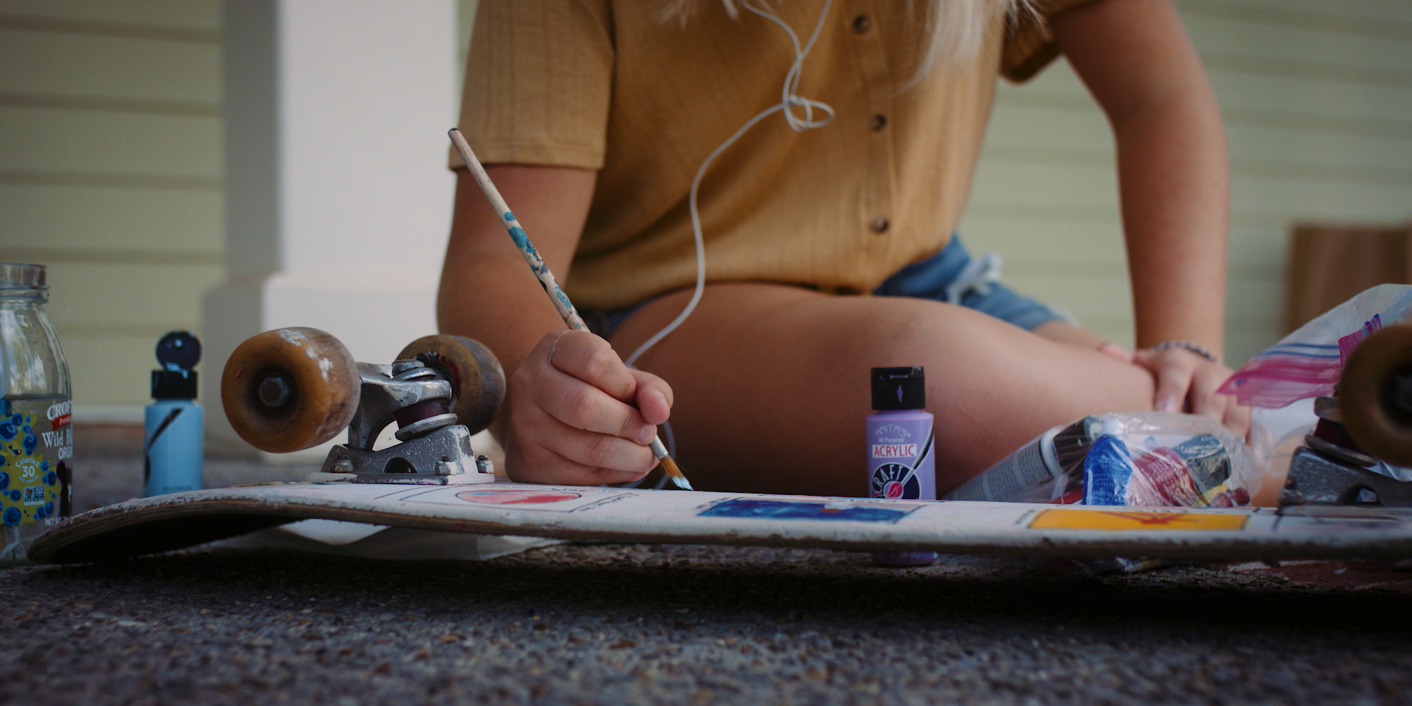 Close-up of a person painting a skateboard with a paintbrush, surrounded by paint bottles and art supplies, sitting on a textured outdoor surface.