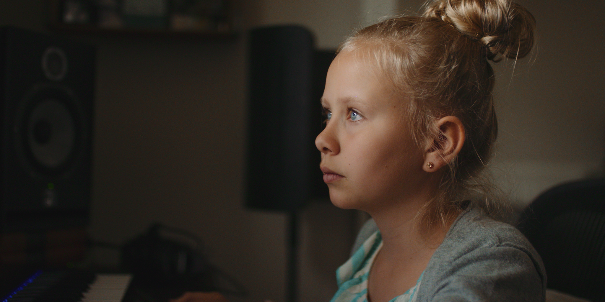 A young girl with blonde hair in a bun, wearing earrings and a striped shirt, sitting in front of a computer monitor, looking intently at the screen.