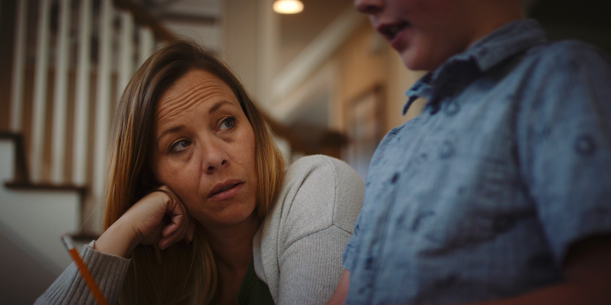 A woman with long hair resting her chin on her hand, looking intently at a person whose face is partially visible, wearing a blue shirt. They are indoors with wooden stairs and shelves in the background.
