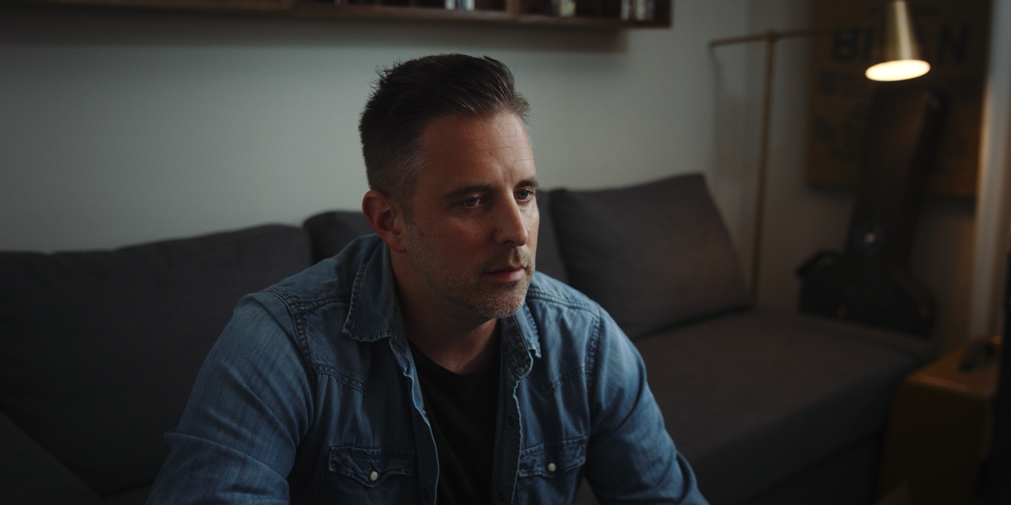 A mid-aged man with dark hair styled back, wearing a denim jacket over a black T-shirt, sitting on a dark gray sofa in a living room, looking at a computer monitor.