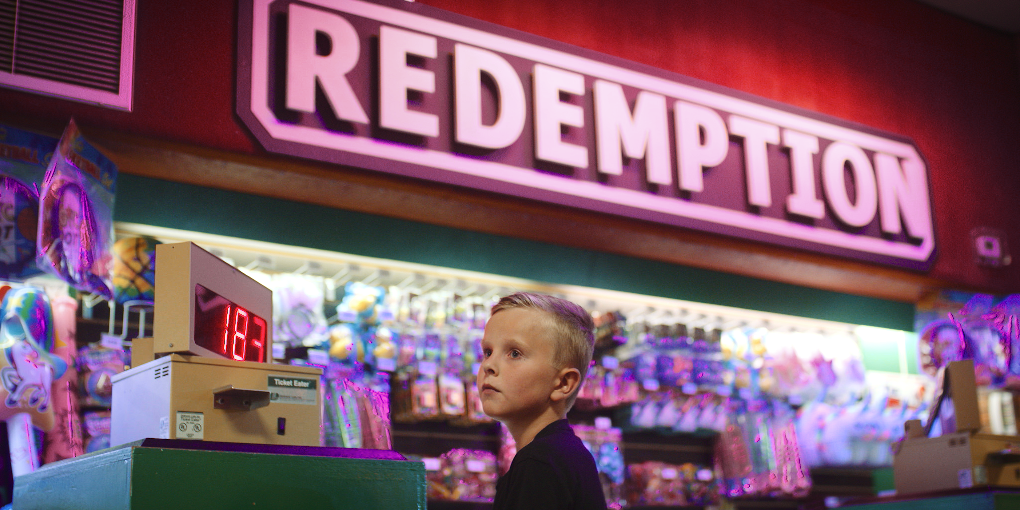 A boy with short blonde hair in a black shirt standing in front of a toy claw game arcade with a pink illuminated sign reading 'Redemption' in the background. The scene is colorful with stuffed toys around.