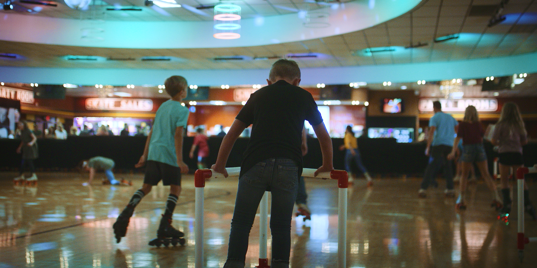 Children roller skating in an indoor skating rink with colorful neon lights and various signs in the background.
