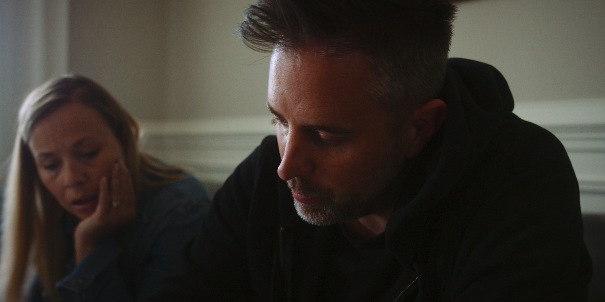 A man with short dark hair and a fade haircut, wearing a black hoodie, appears to be in distress or deep thought. Behind him, a woman with blonde hair, wearing a gray jacket, looks concerned and has her hand near her face, possibly touching her cheek or jaw. The scene seems to be indoors with neutral-colored walls.