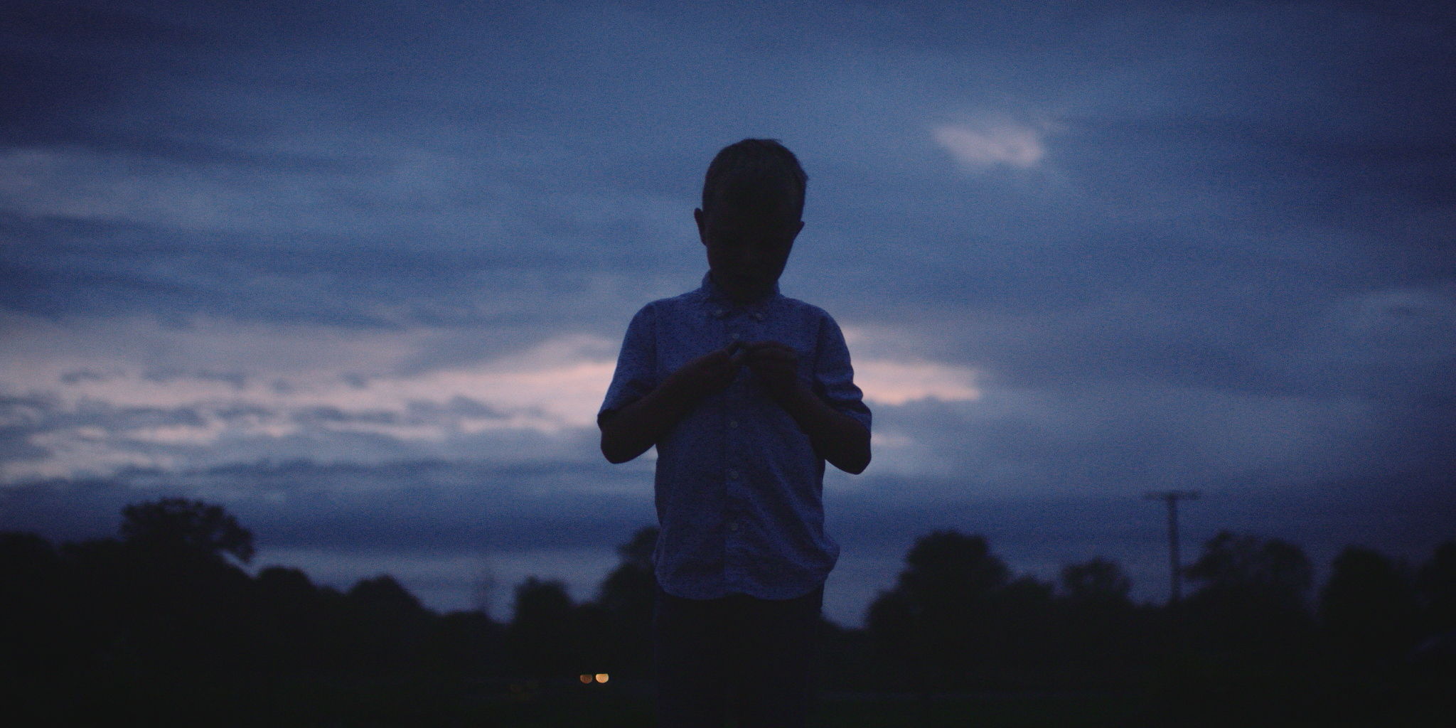 Silhouette of a boy standing outdoors at dusk, holding an object, with a cloudy sky in the background and trees along the horizon.