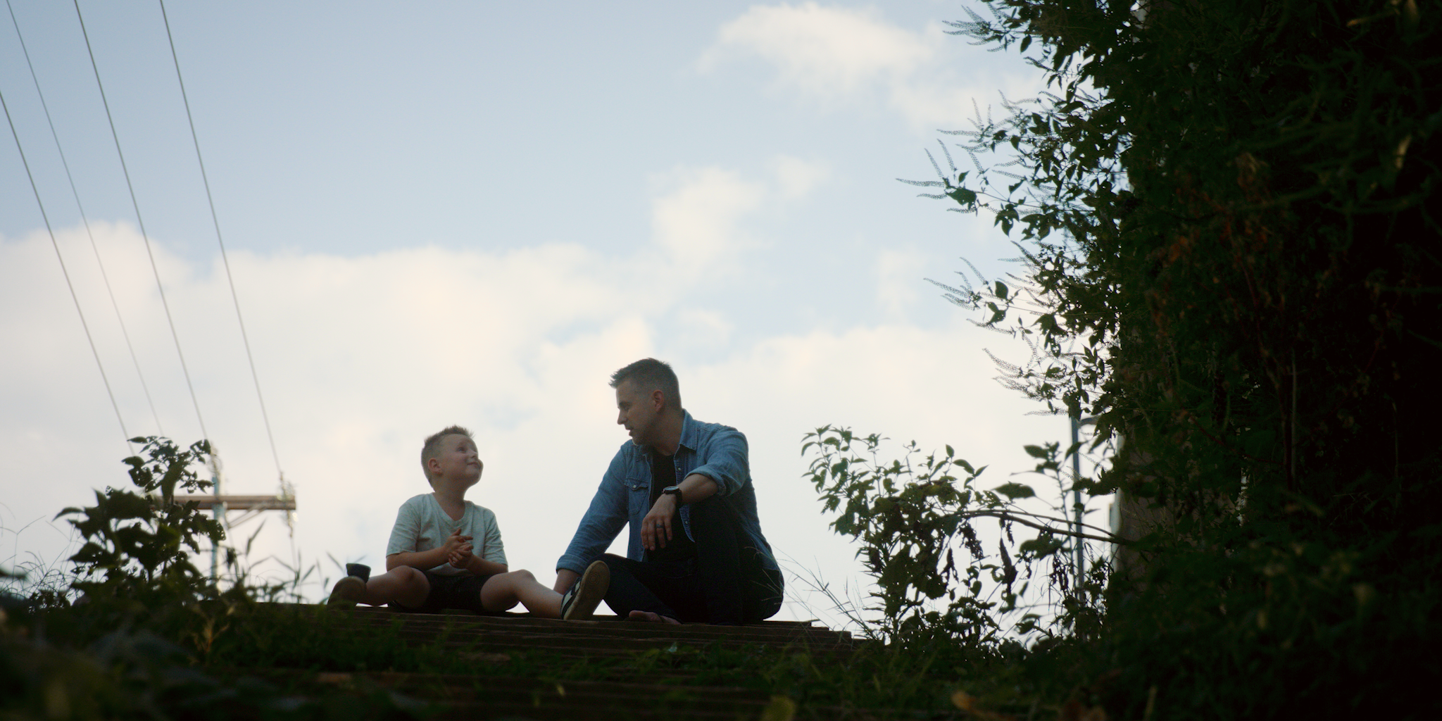 A man and a boy sitting together outdoors, engaging in conversation during daylight hours, with trees and power lines in the background.