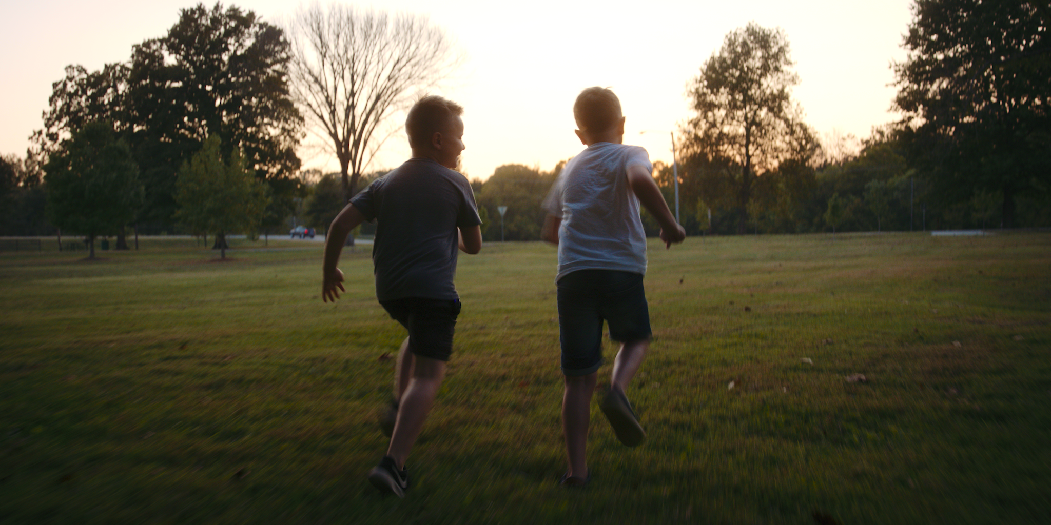 Two children run across a grassy field with trees and a sunset sky in the background.