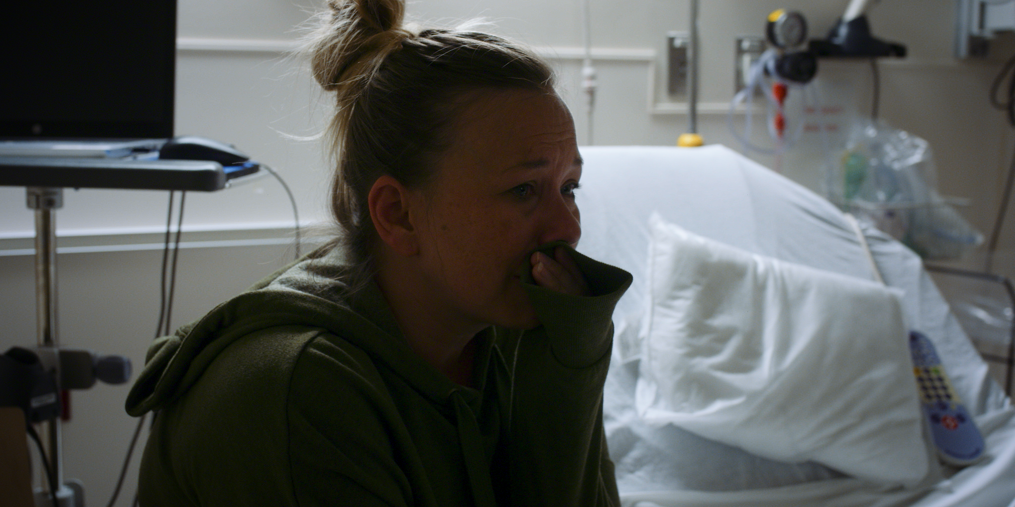 A woman sitting in a hospital room, holding a tissue to her nose, with medical equipment and a hospital bed in the background.