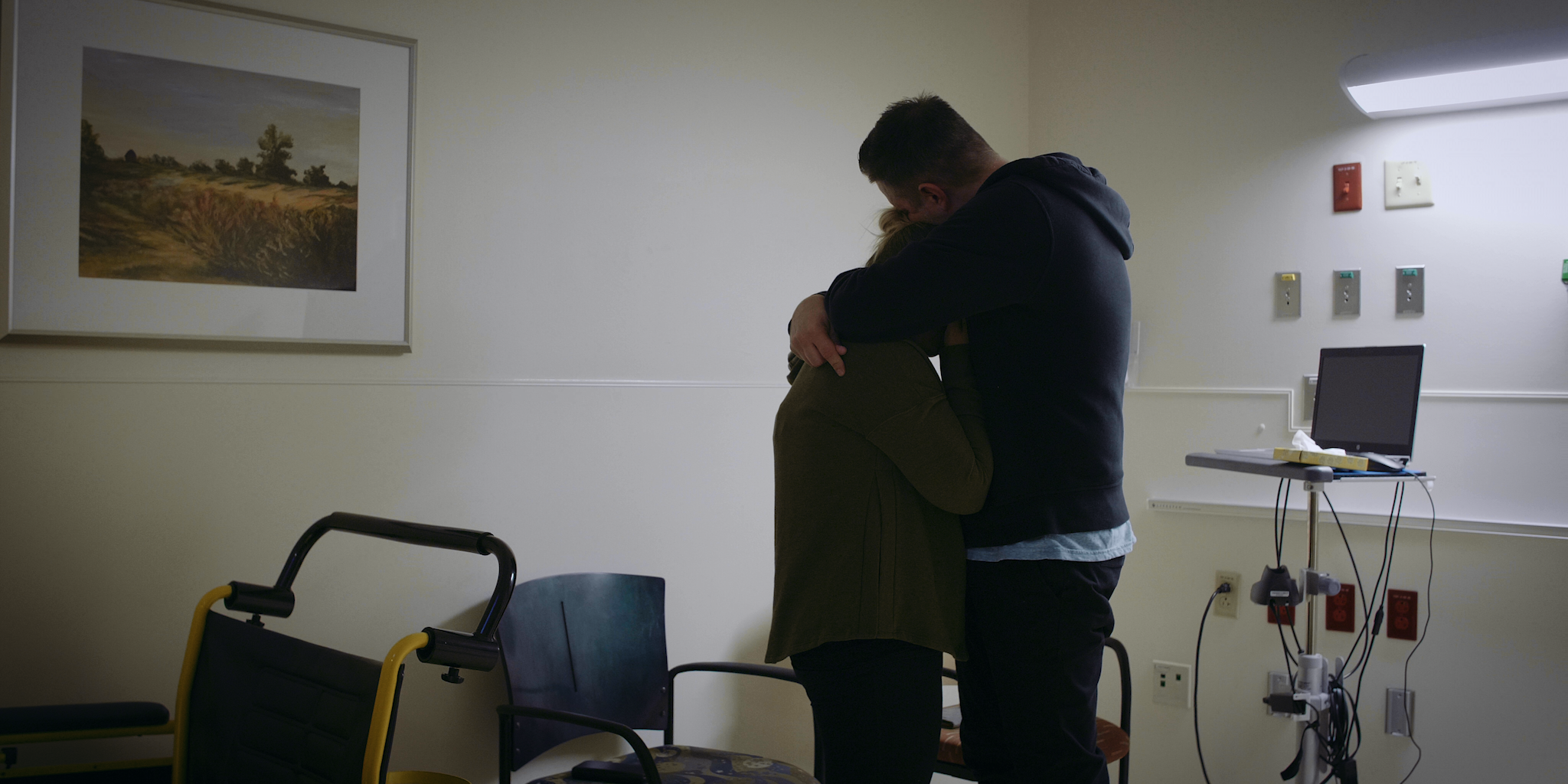 A person hugging a woman in a hospital room with chairs, a framed painting, and medical equipment in the background.