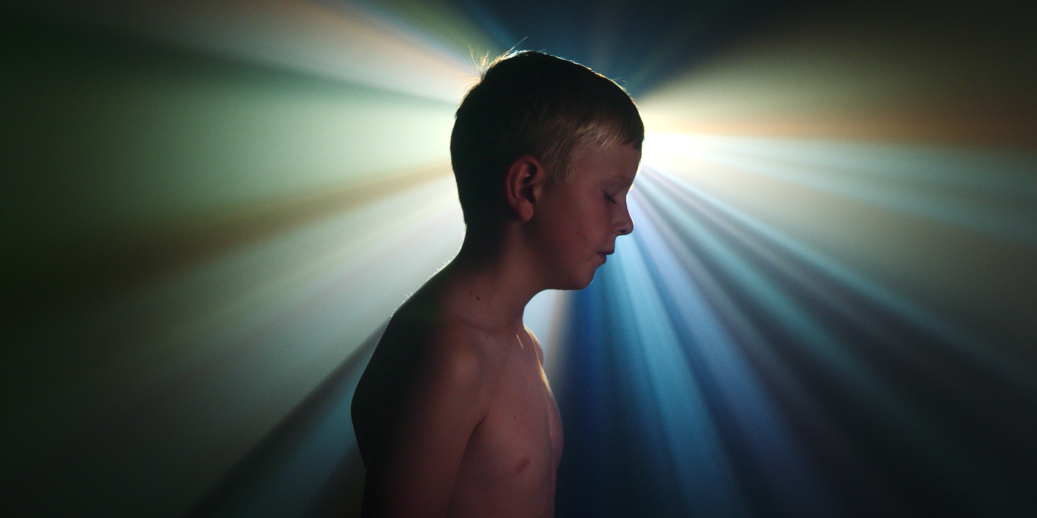 A young boy with short hair and closed eyes, standing shirtless with light rays and colorful glow behind him.