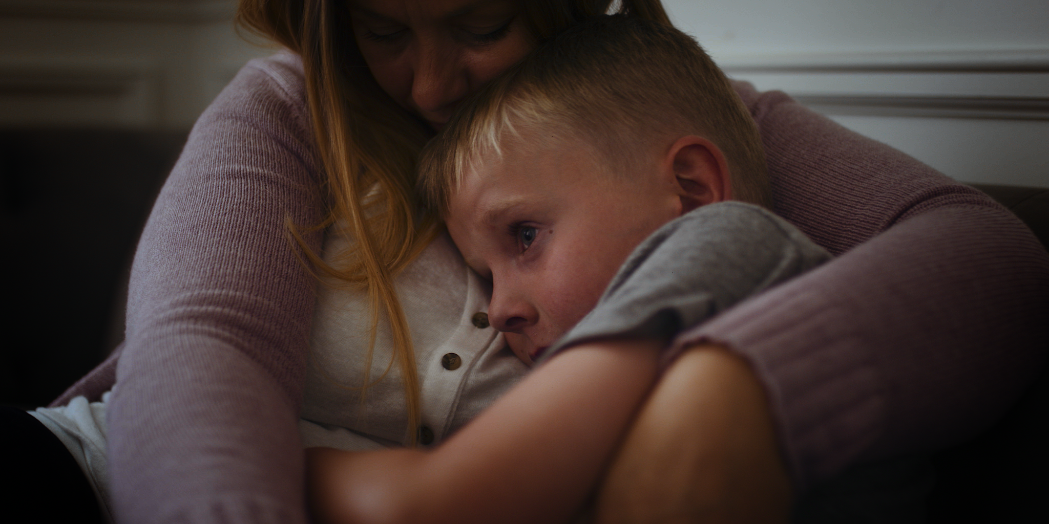 A woman and a young boy embrace each other, with the woman gently resting her face on the boy's head while he looks pensively into the distance.