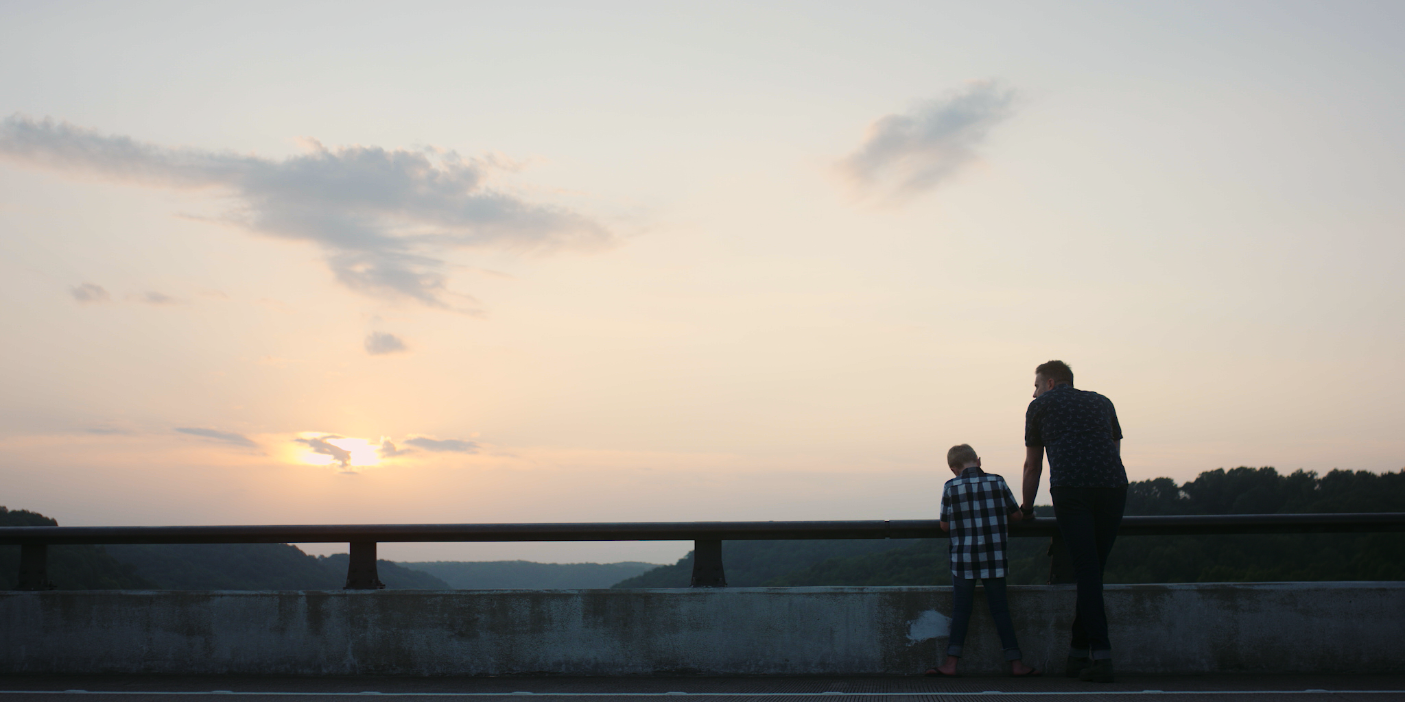 A man and a boy standing on a bridge, watching the sunset over a river with trees in the distance.