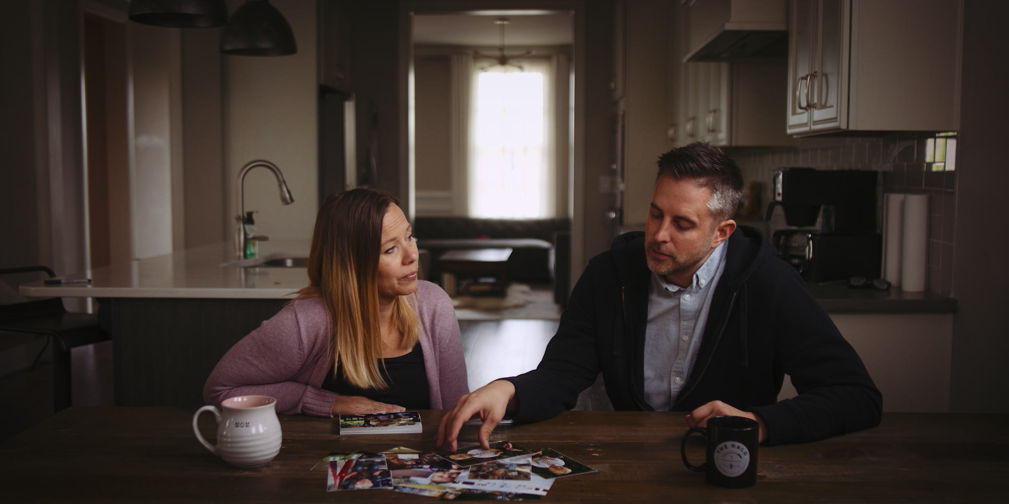 A man and woman sitting at a dining table in a kitchen, looking at photographs spread out on the table, with a coffee mug and a mug labeled 'Mom' in the foreground.