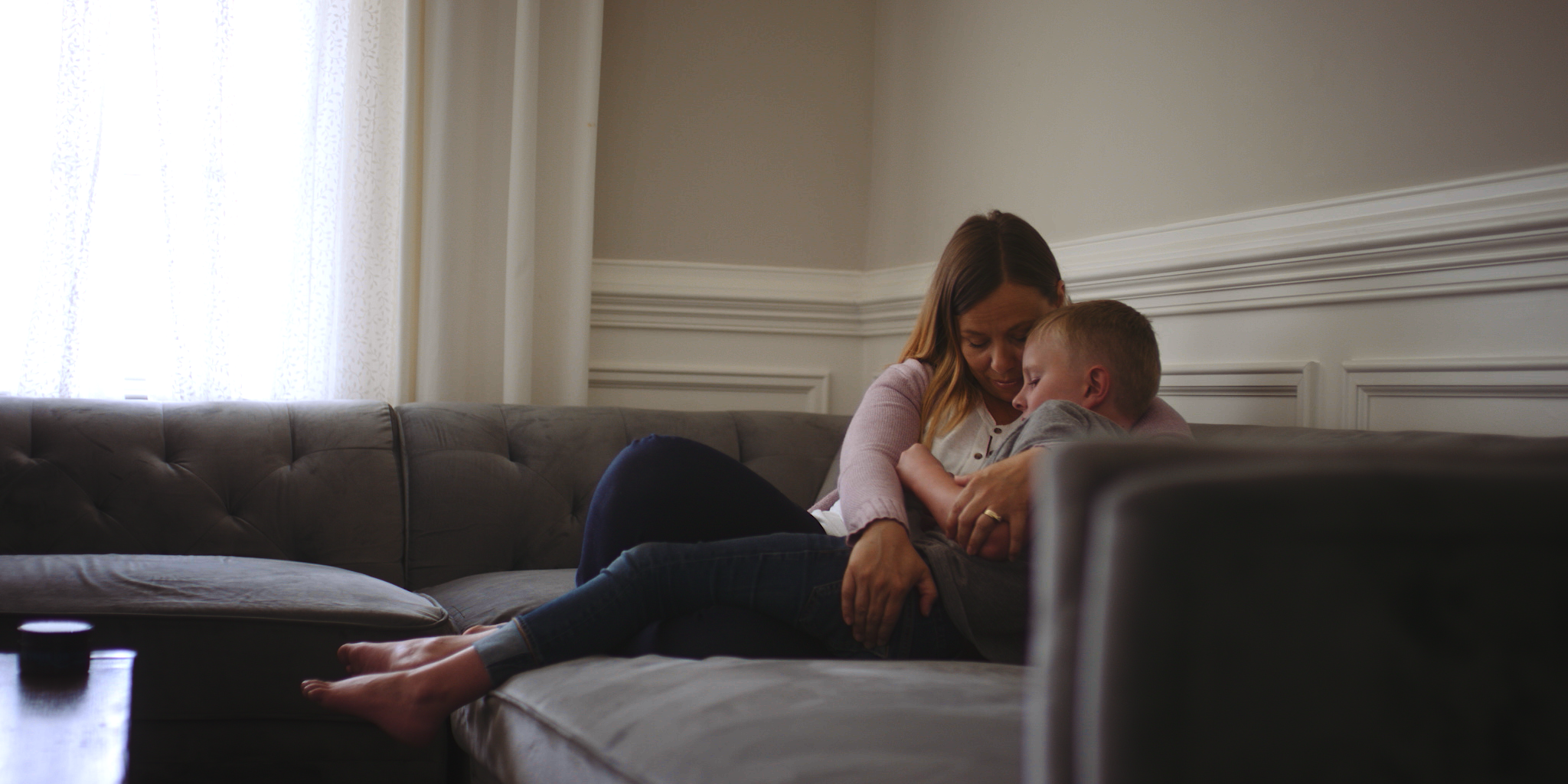 A woman and a young boy sitting on a gray couch in a well-lit living room, sharing an intimate moment with their heads resting on each other's shoulders.