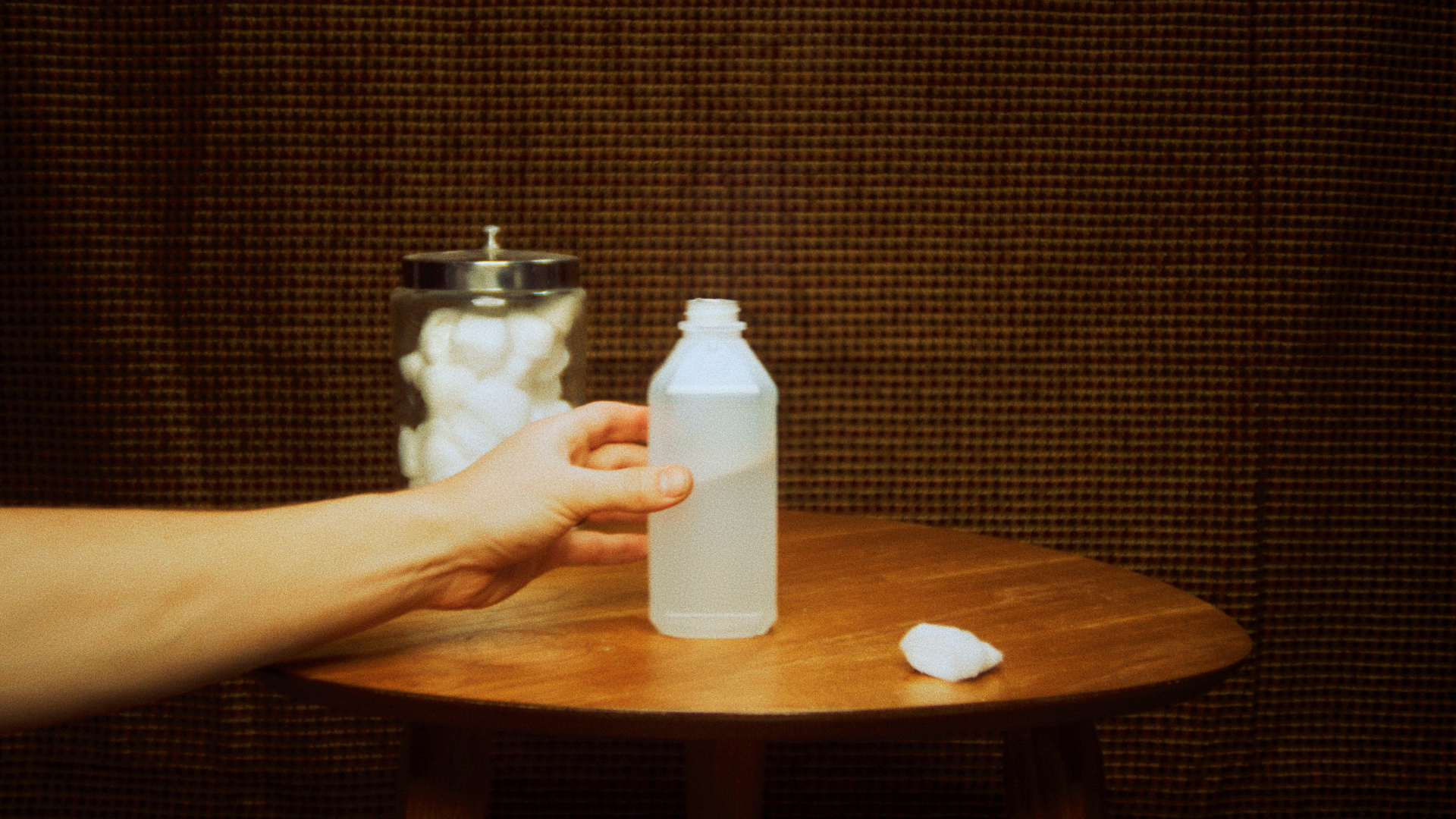 A hand holding a small white bottle in front of a jar filled with cotton balls on a wooden table.