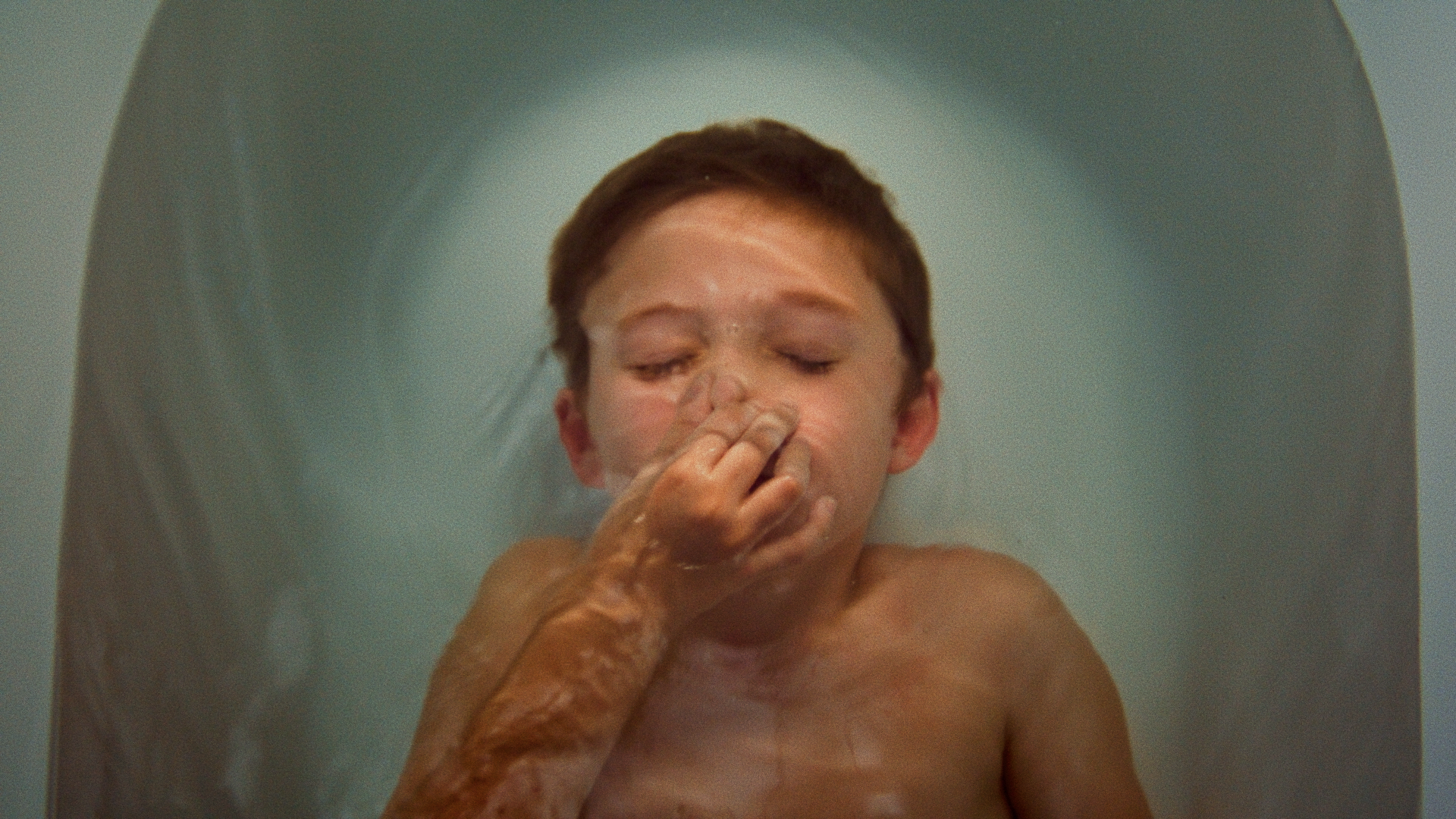 A young boy with closed eyes in a bathtub, a person’s hand pinching his nose, filling the scene with water.