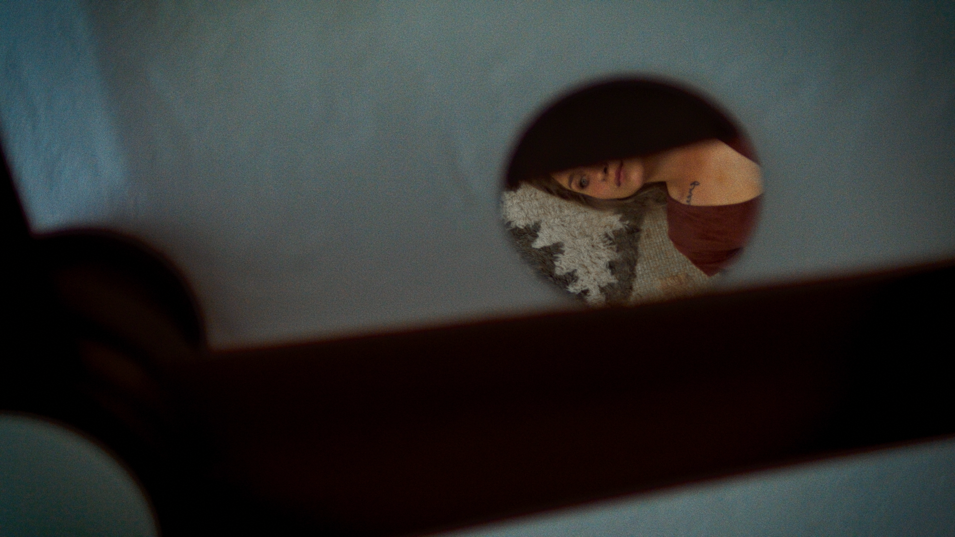 A woman with brunette hair, wearing a reddish-brown top, seen through a small circular hole from an overhead perspective, reflecting on a mirror or window.