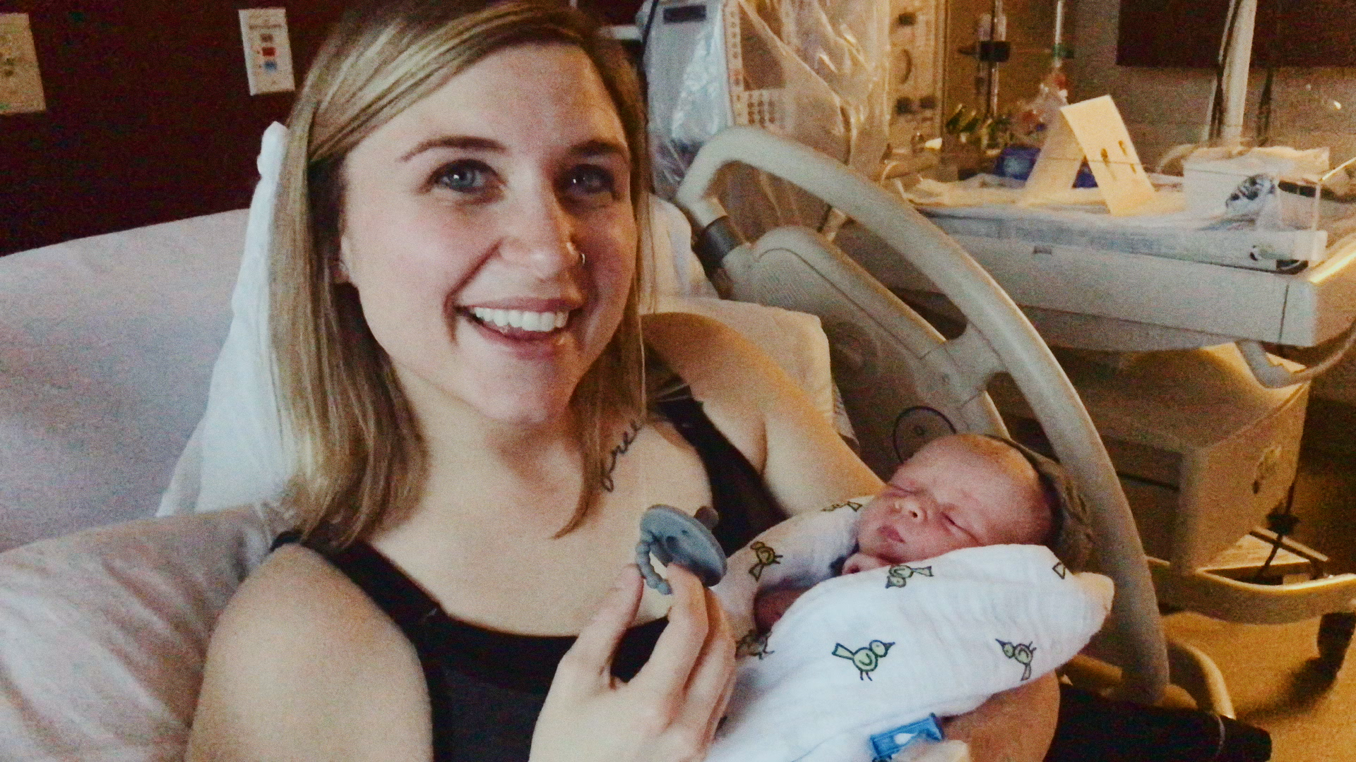 A woman with blonde hair and a nose ring smiling in a hospital room, holding a newborn baby wrapped in a blanket.
