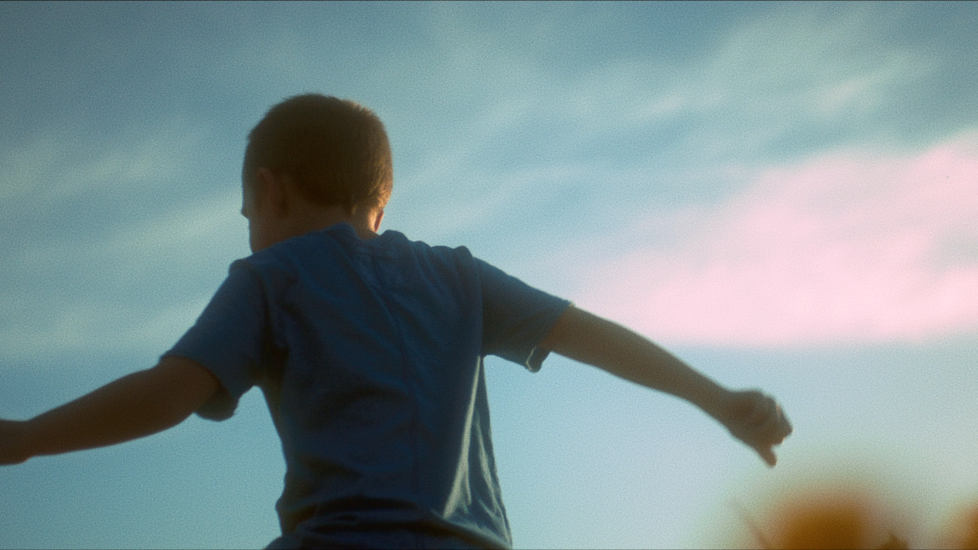 A boy with short hair wearing a blue shirt, standing outdoors with arms outstretched, under a bright blue sky.