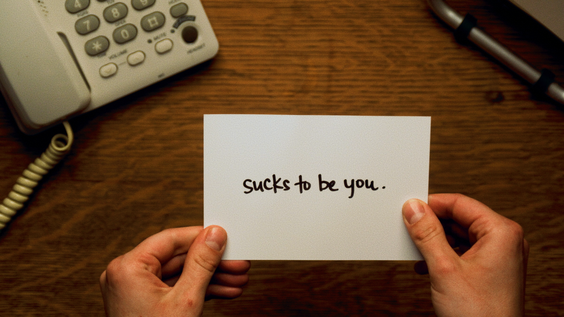 A person holding a white card with the handwritten message "sucks to be you." in black ink. In the background, there is a desk with a white landline phone and a pen.