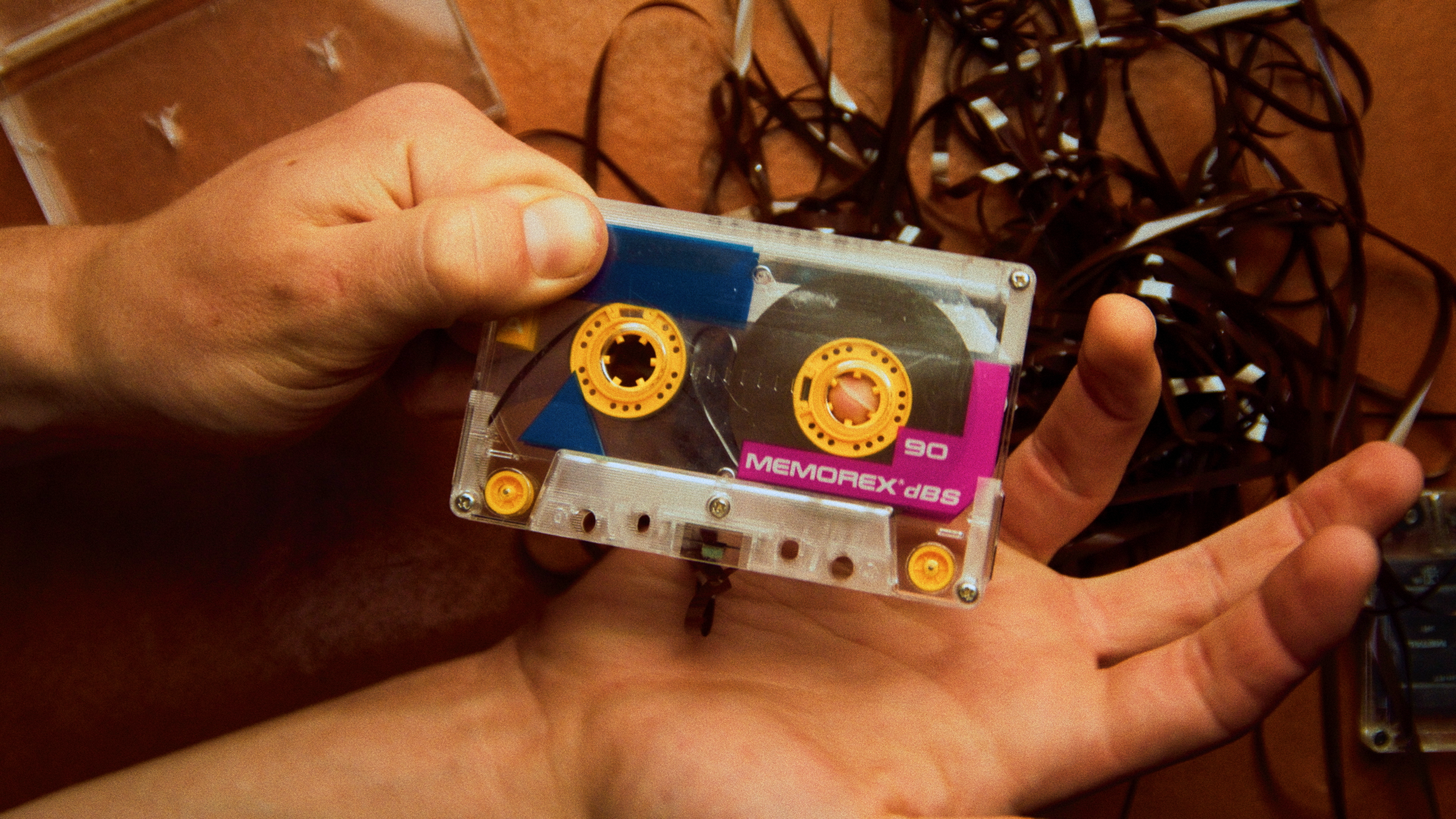 A person holding a cassette tape labeled 'MEMOREX dBS 90' in front of a tangled pile of dark cables on a wooden surface.