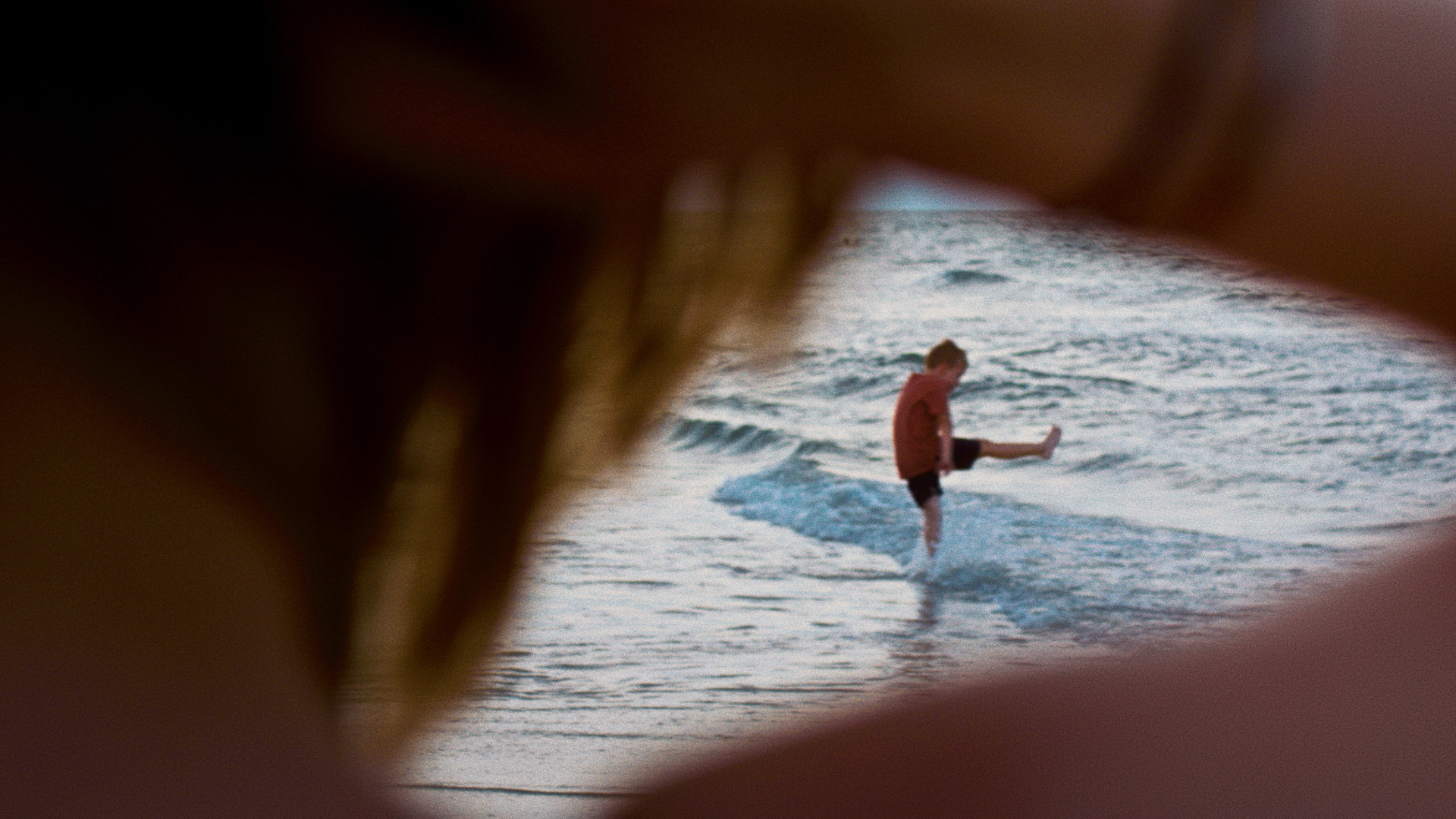 A young boy wearing a red shirt and black shorts standing in shallow ocean water, kicking his leg in the air while being viewed through a blurry, foreground object on the beach.