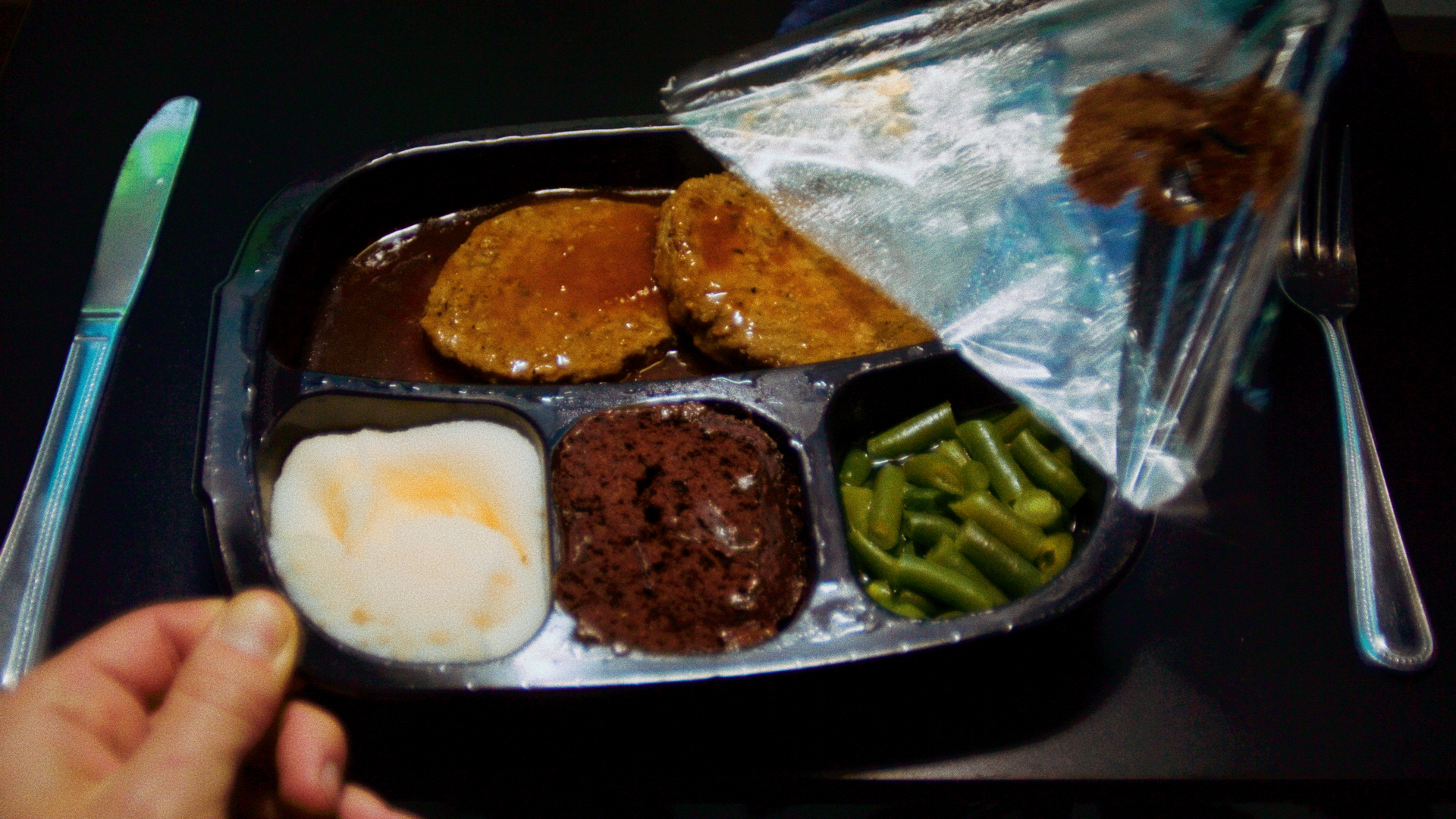 A TV dinner being unpeeled. A person's hand is holding the tray, and utensils are placed on either side of the tray.