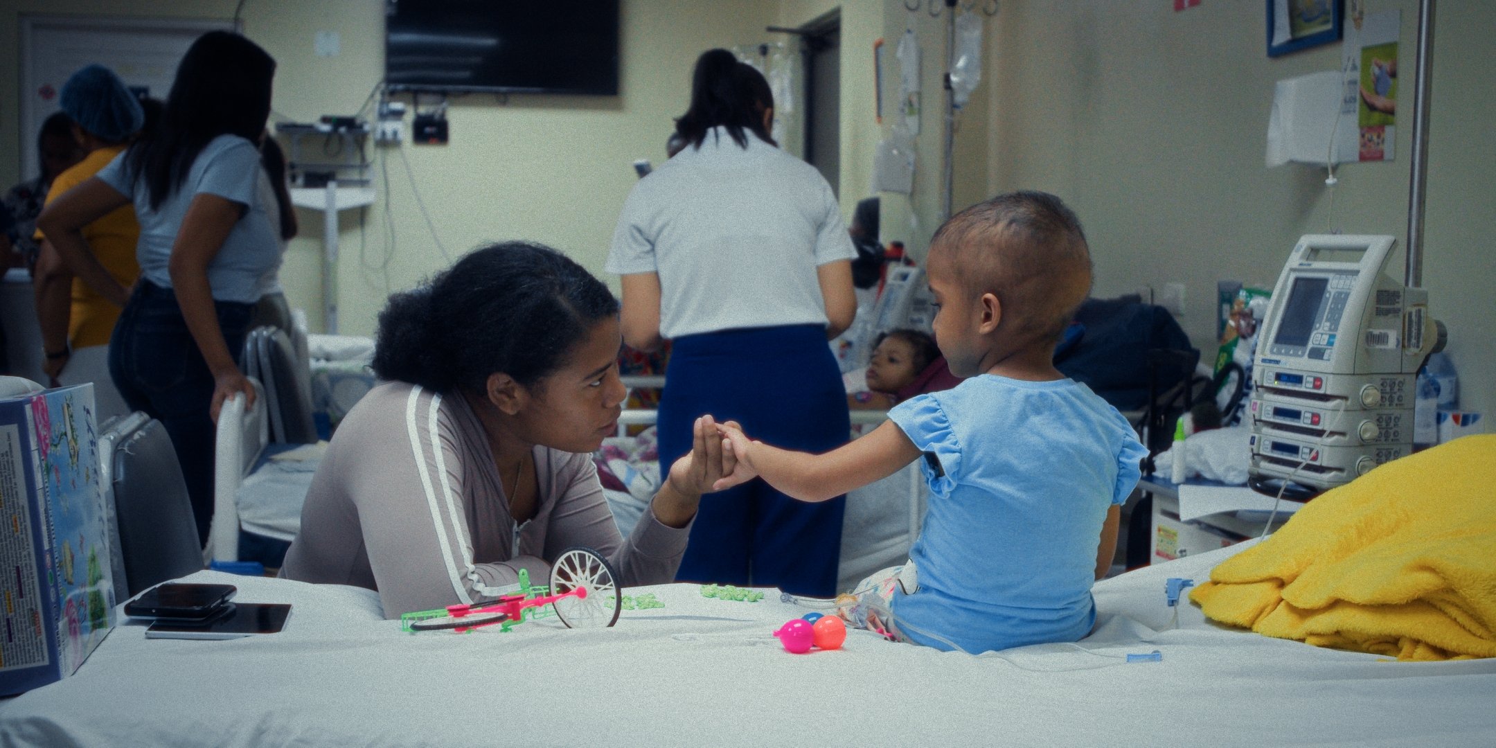 A healthcare setting with a young boy and a woman, possibly his mother, holding hands and looking at each other. Other medical staff and children are present, with hospital equipment and toys on the table.