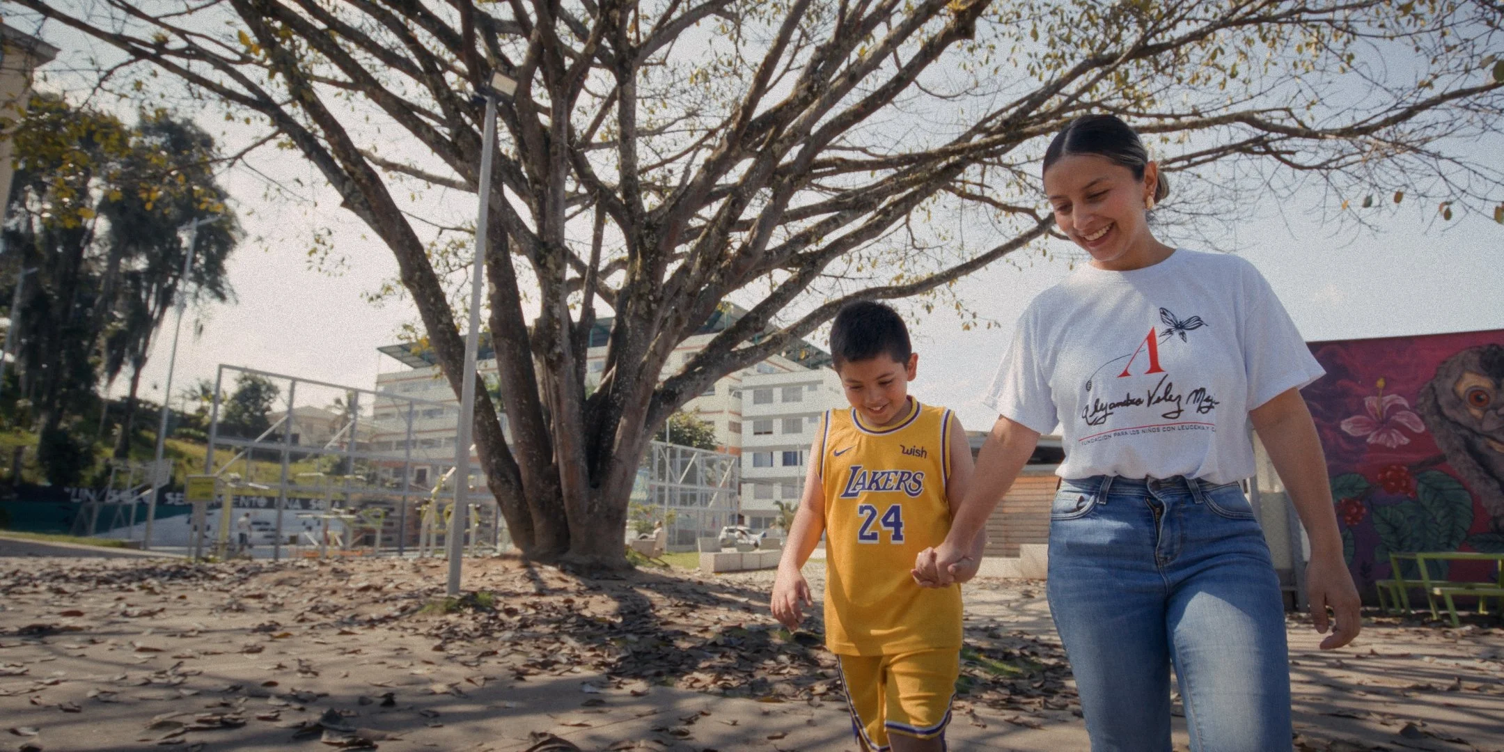 A woman and a boy walking hand in hand outdoors on a tree-lined pathway, with fallen leaves on the ground and a building in the background.