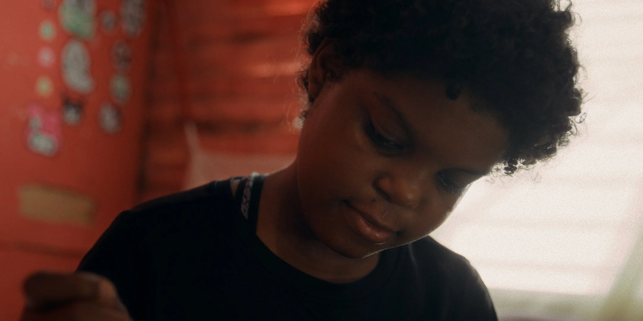 A young child with curly hair looking down, holding an orange object with assorted stickers on it.