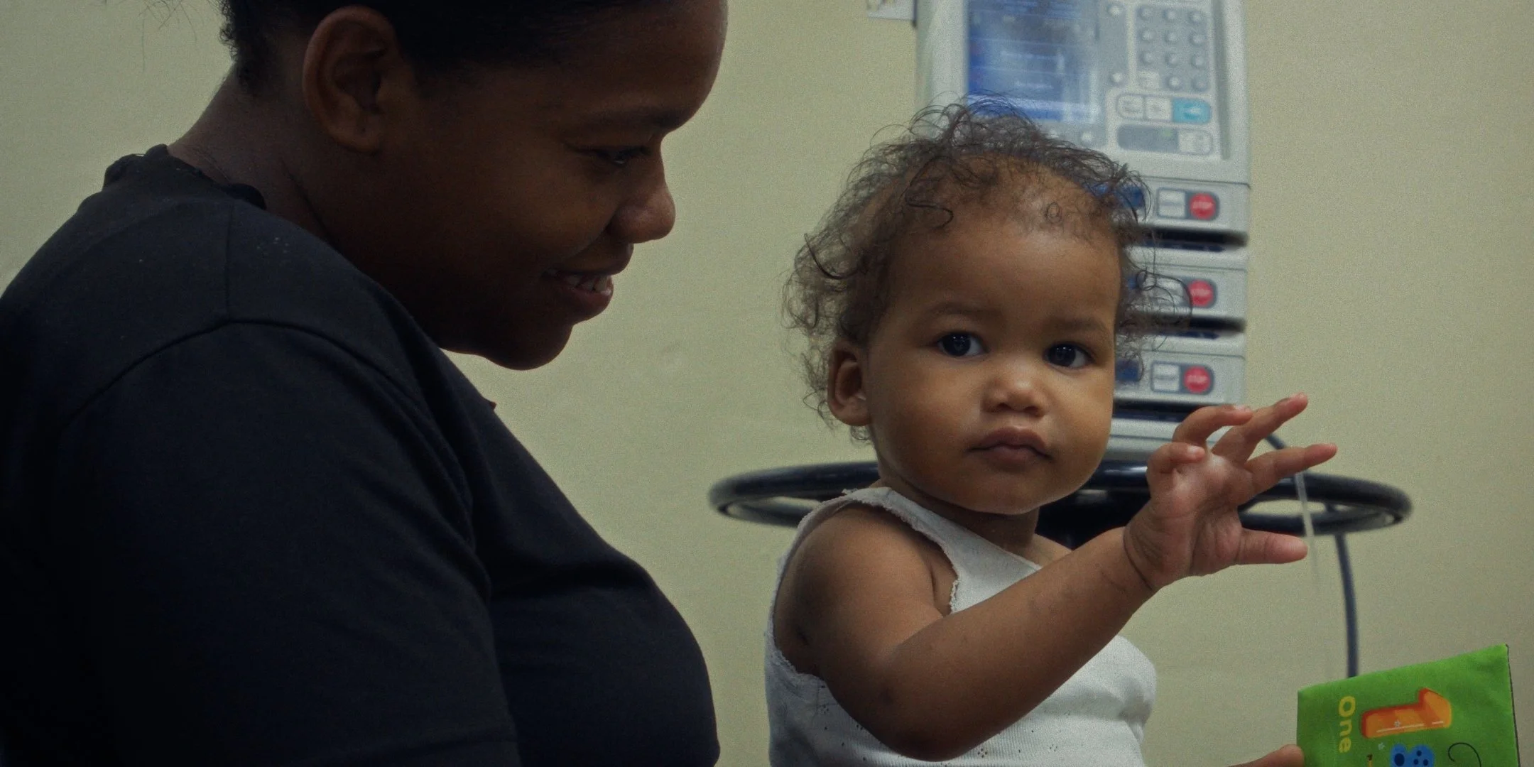 A woman and a young girl sitting at a table, with the girl holding a colorful book and the woman smiling, in a room with medical equipment visible in the background.