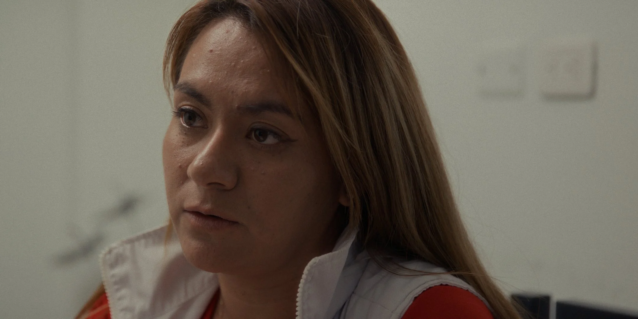 Close-up of a woman with long reddish hair, wearing a white zip-up shirt with red accents, looking to the side with a serious expression.