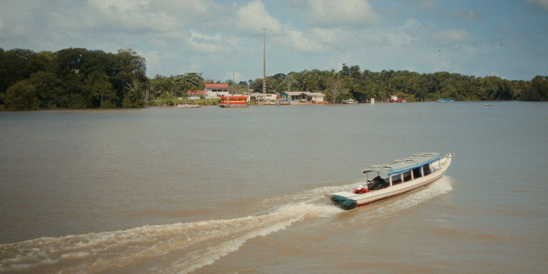 A boat traveling on a river near a settlement with trees and buildings, under a partly cloudy sky.