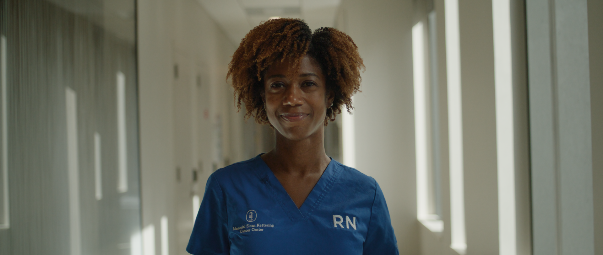 A smiling woman with curly hair wearing blue medical scrubs with the words "Memorial Sloan Kettering Cancer Center" and "RN" on it, standing in a hospital corridor with windows on the right.