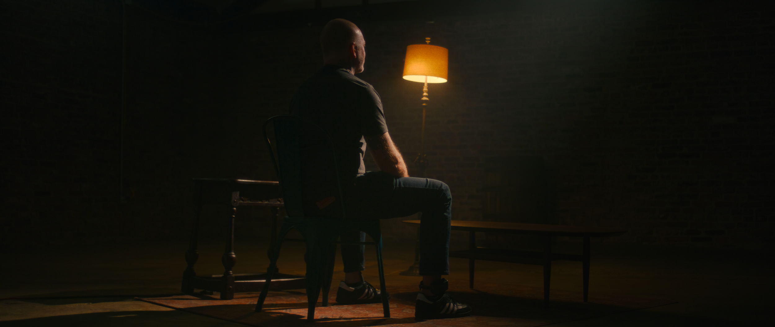 A man sitting on a chair in a dimly lit room, facing side profile. There is a warm-colored floor lamp behind him illuminating the space, with a small table in front of him and a couch to his side.