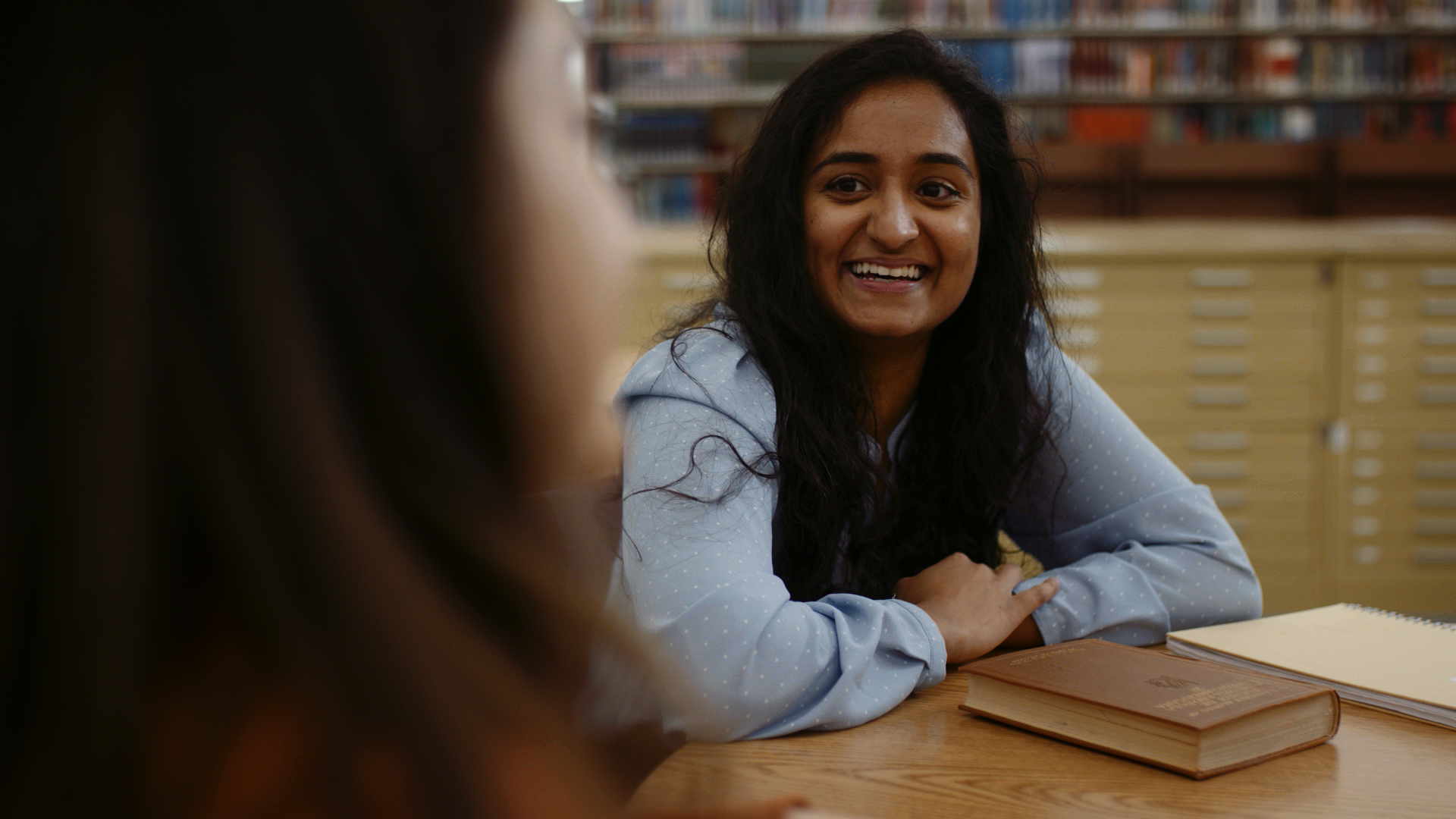 A woman with long dark hair smiling and talking to someone across a wooden table in a library, with books visible in the background.