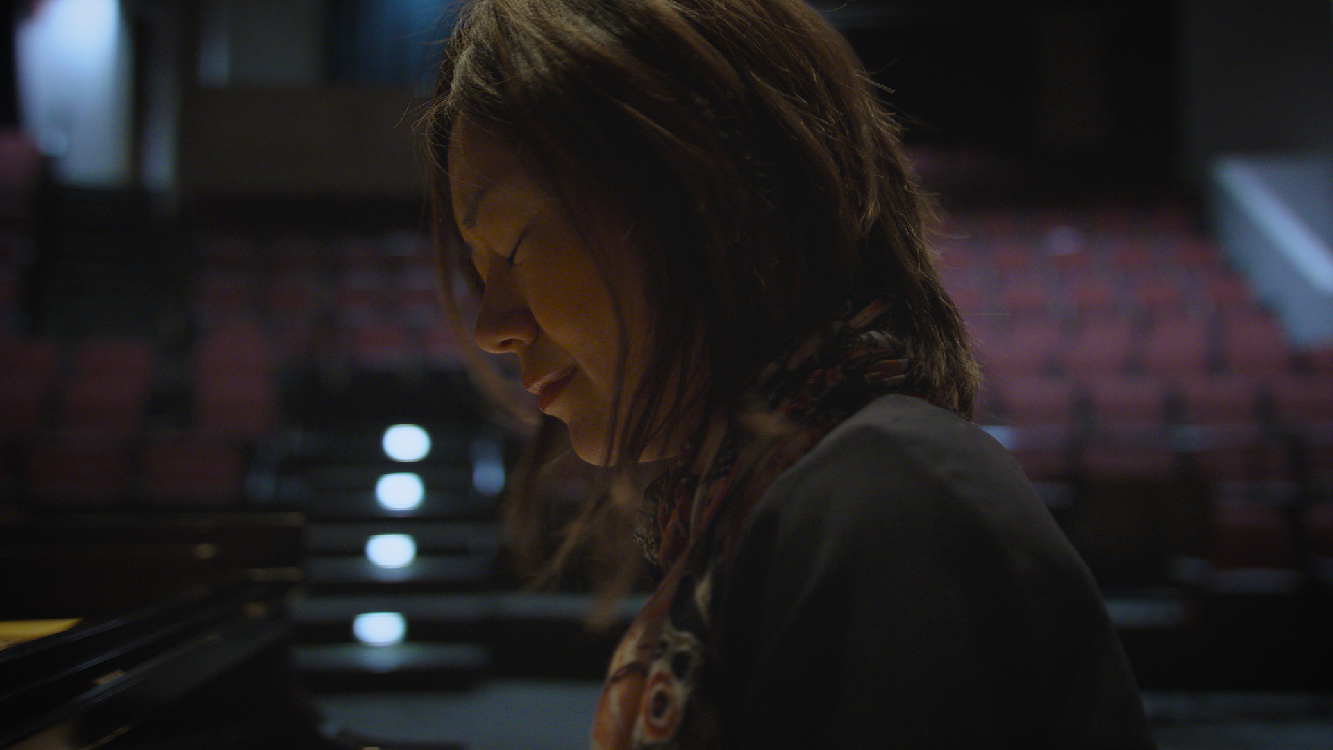 A woman with brown hair, wearing a patterned scarf, is playing a piano in a dimly lit auditorium.