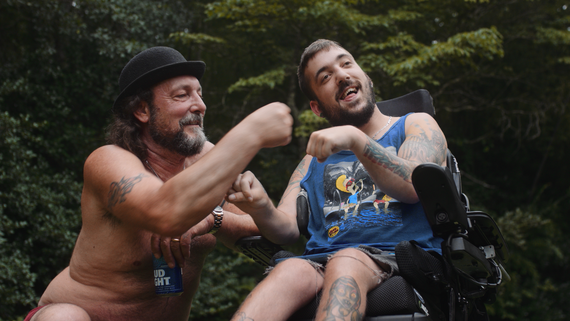 A shirtless man with tattoos and a black hat making fists with a bearded man in a wheelchair, also with tattoos, wearing a blue tank top with a graphic design, outdoors surrounded by trees.