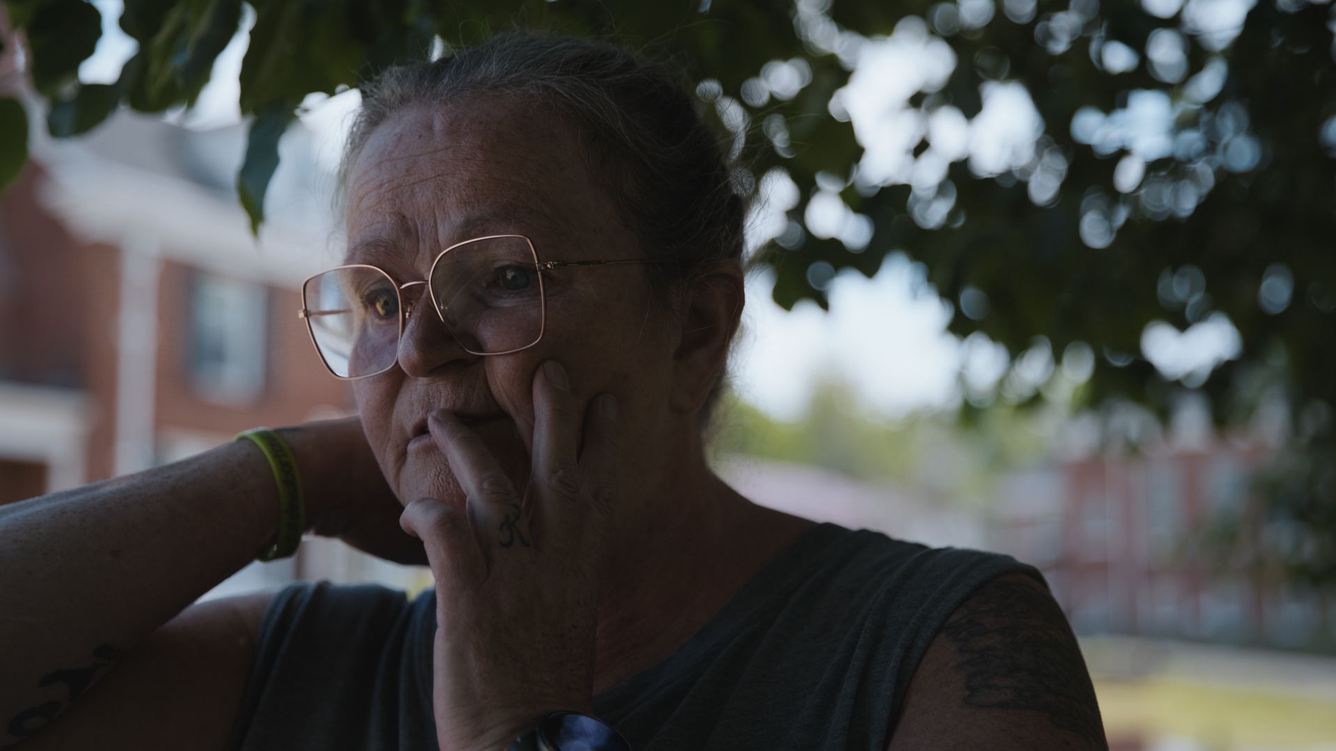A woman with glasses, dark hair tied back, and tattoos on her arm, touching her face and looking downward outside with trees and a building in the background.