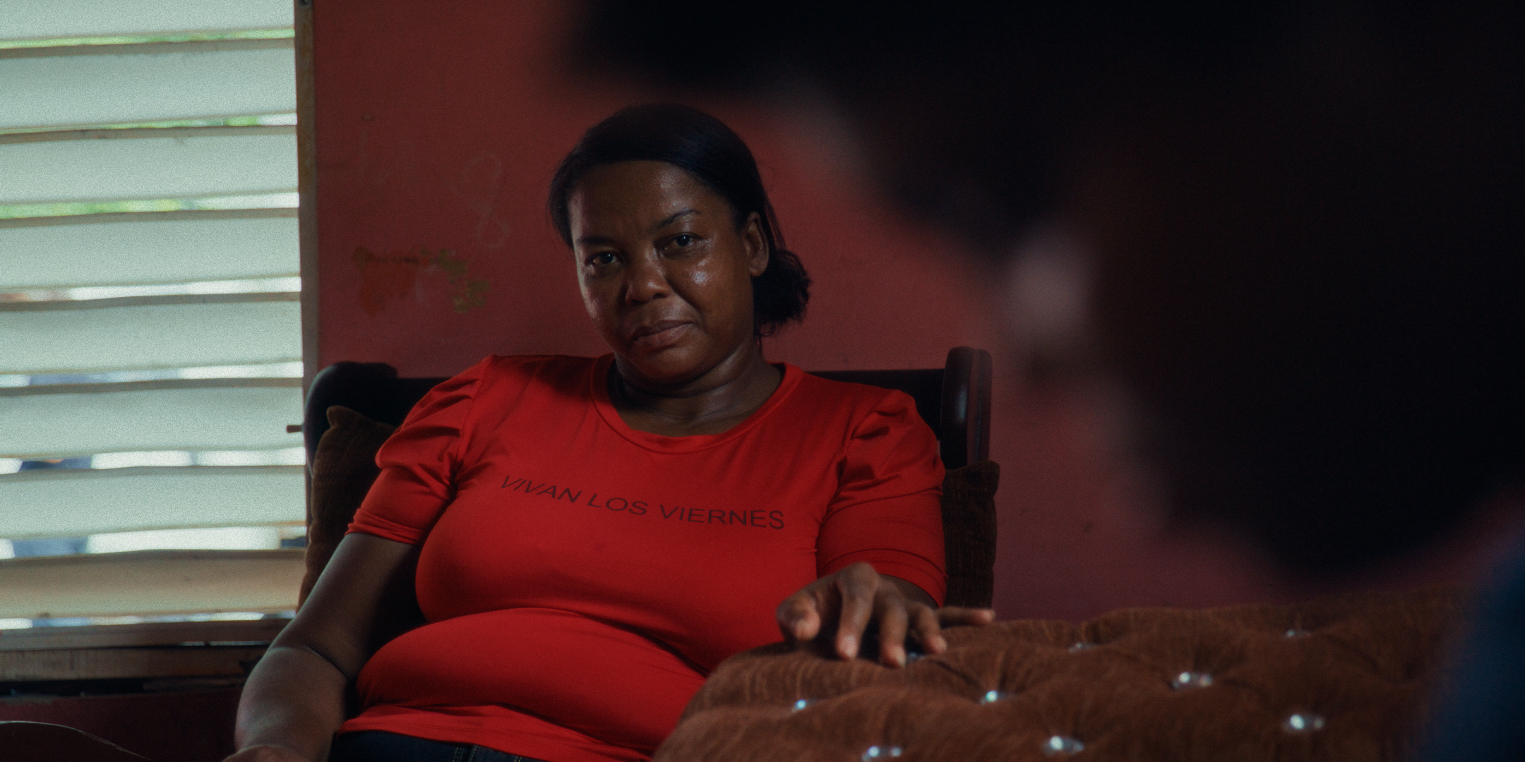 A woman with dark hair in a ponytail sitting on a couch in a room with striped blinds and a red wall. She is wearing a red shirt and looking directly at the camera.