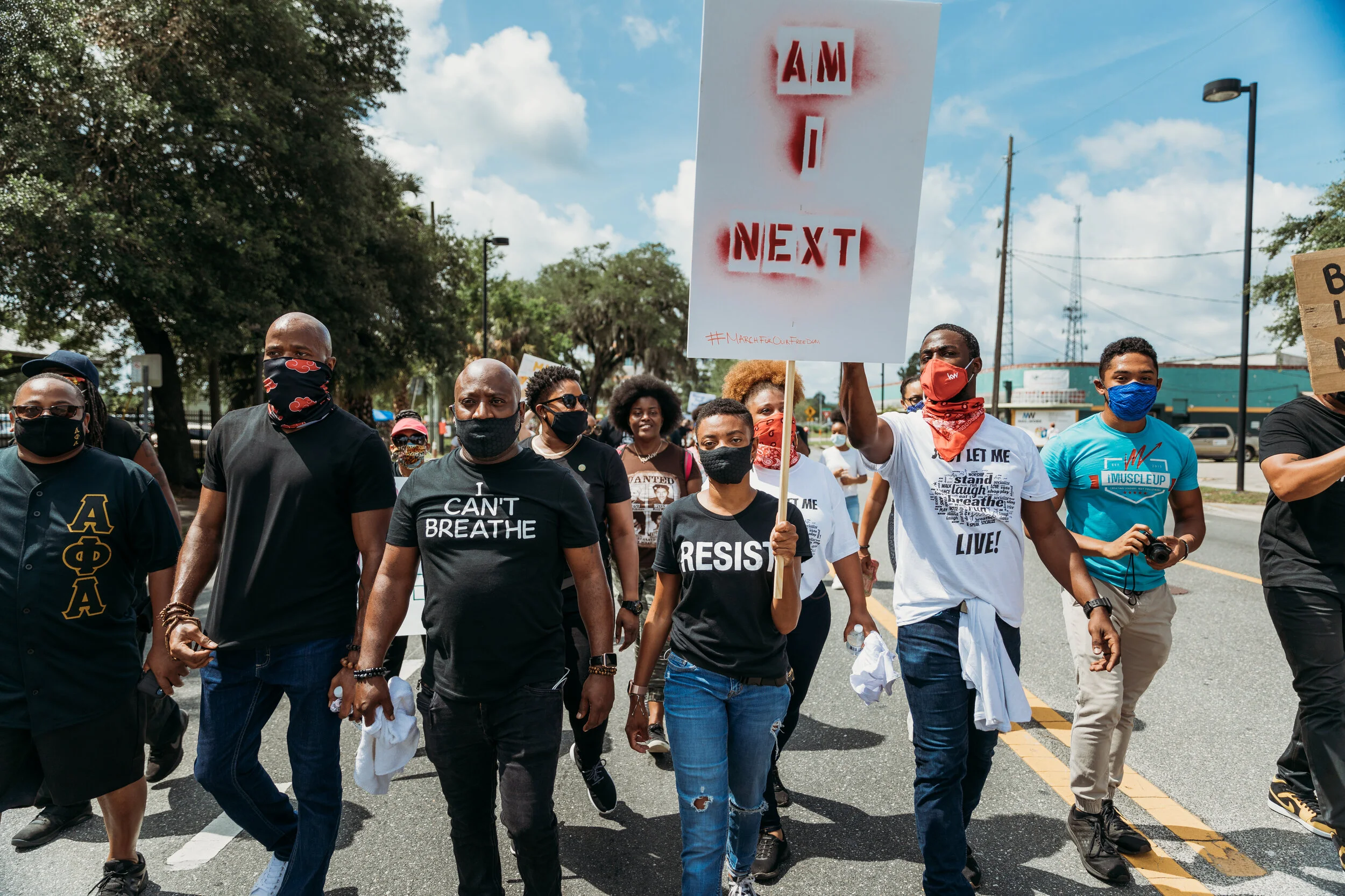 A diverse group of people participating in a protest march, many wearing black face masks, holding signs including one that reads 'AM I NEXT.'